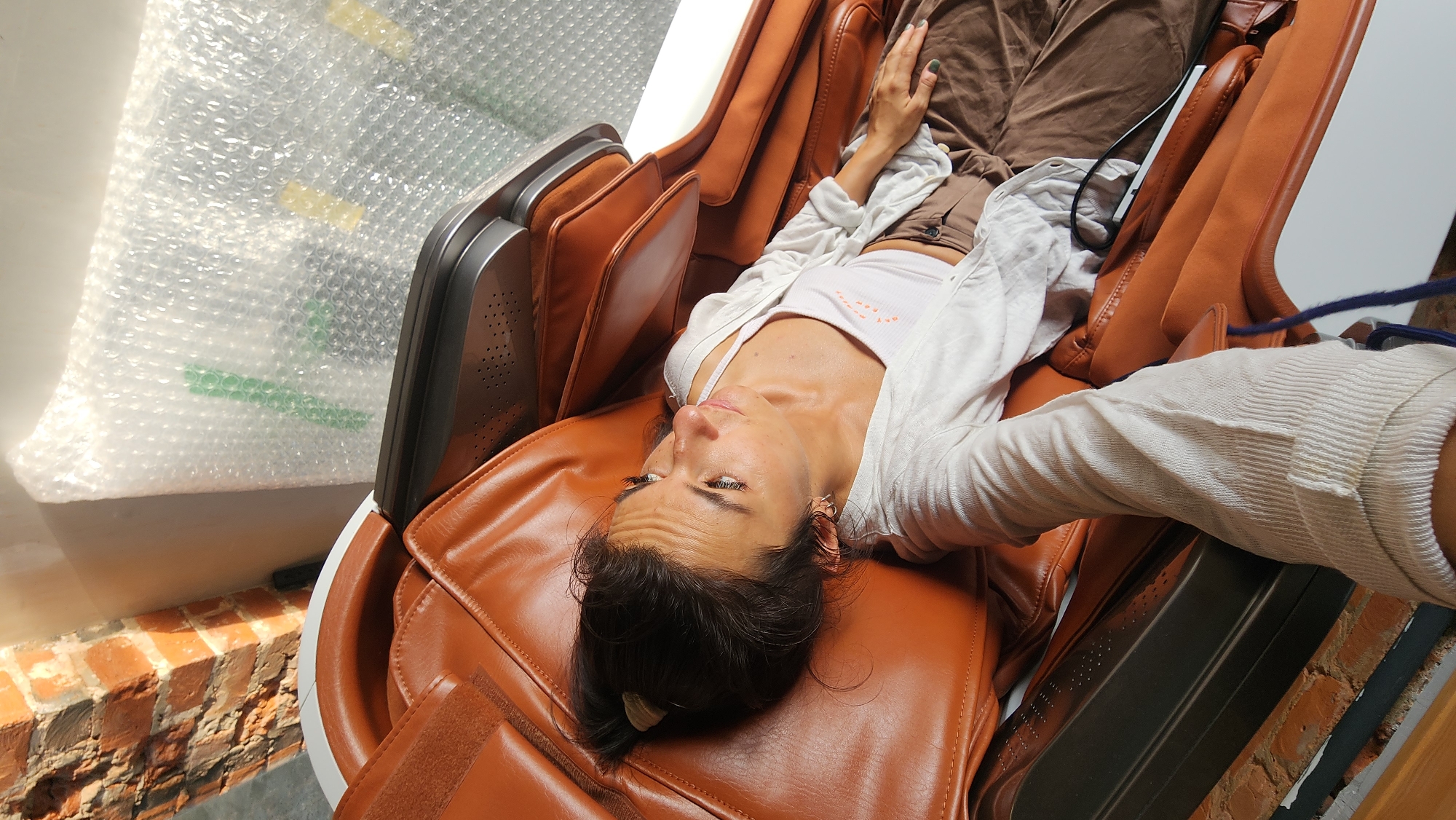 A woman with dark hair pulled back in a ponytail reclines in a brown massage chair. She is wearing a white tank top and a light-colored, long-sleeved open shirt over it, along with brown pants. Her head is turned to her right, and her eyes are looking upwards. The background shows a brick wall on the left and a white surface covered in bubble wrap on the right. The overall scene appears to be indoors, possibly in a commercial space or a private residence in Moscow, Russia.