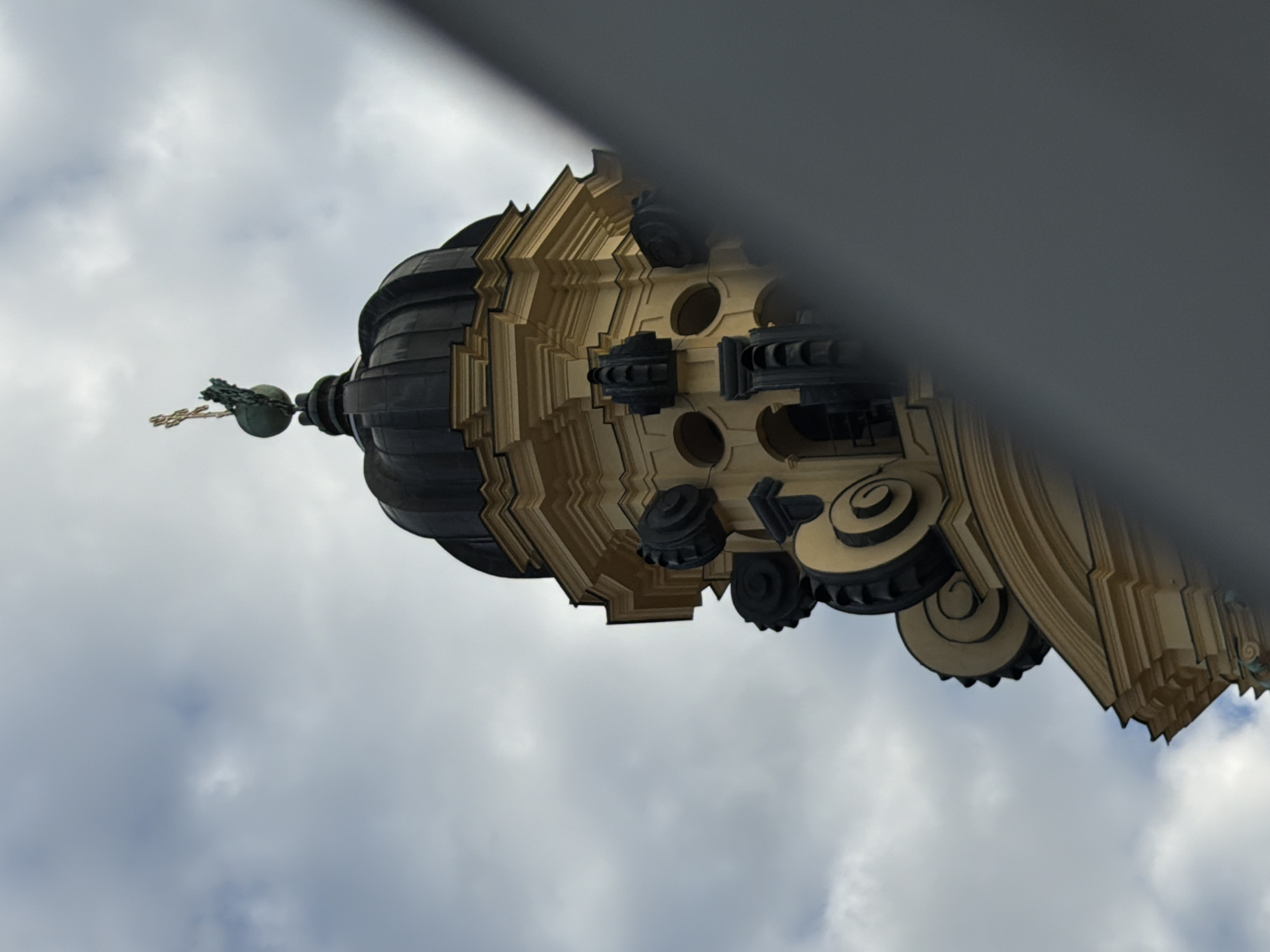 A baroque-style dome, painted yellow with black decorative elements, rises into a cloudy sky. A golden cross topped with a sphere sits at the apex of the dome. The building appears to be a church, likely part of the Frauenkirche in Munich, Germany. The lower right portion of the image is obscured by a blurred foreground element, possibly a fence or wall.