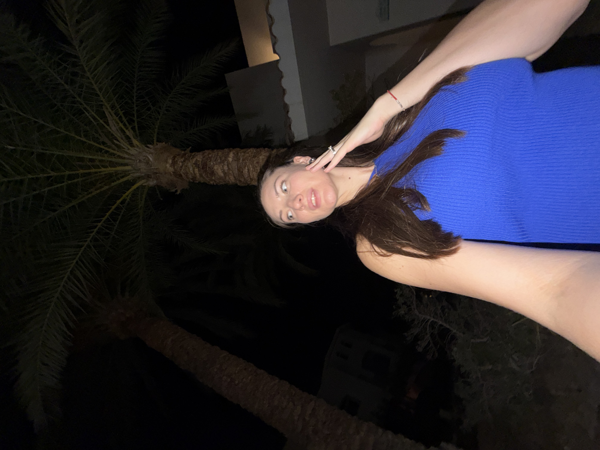 A woman is captured in a low-angle shot at night, likely in the city of Sharm El Sheikh, Egypt, with two large palm trees framing the scene. The woman, with long brown hair, is wearing a bright blue ribbed top. Her right hand is raised to her face, touching her cheek, and she is looking away from the camera to her left. She is wearing a ring on her left ring finger and a thin red bracelet on her right wrist. Behind her, a building with a tiled roof is visible, illuminated by a warm light. The overall atmosphere suggests an outdoor setting at a resort or hotel, possibly during a vacation.