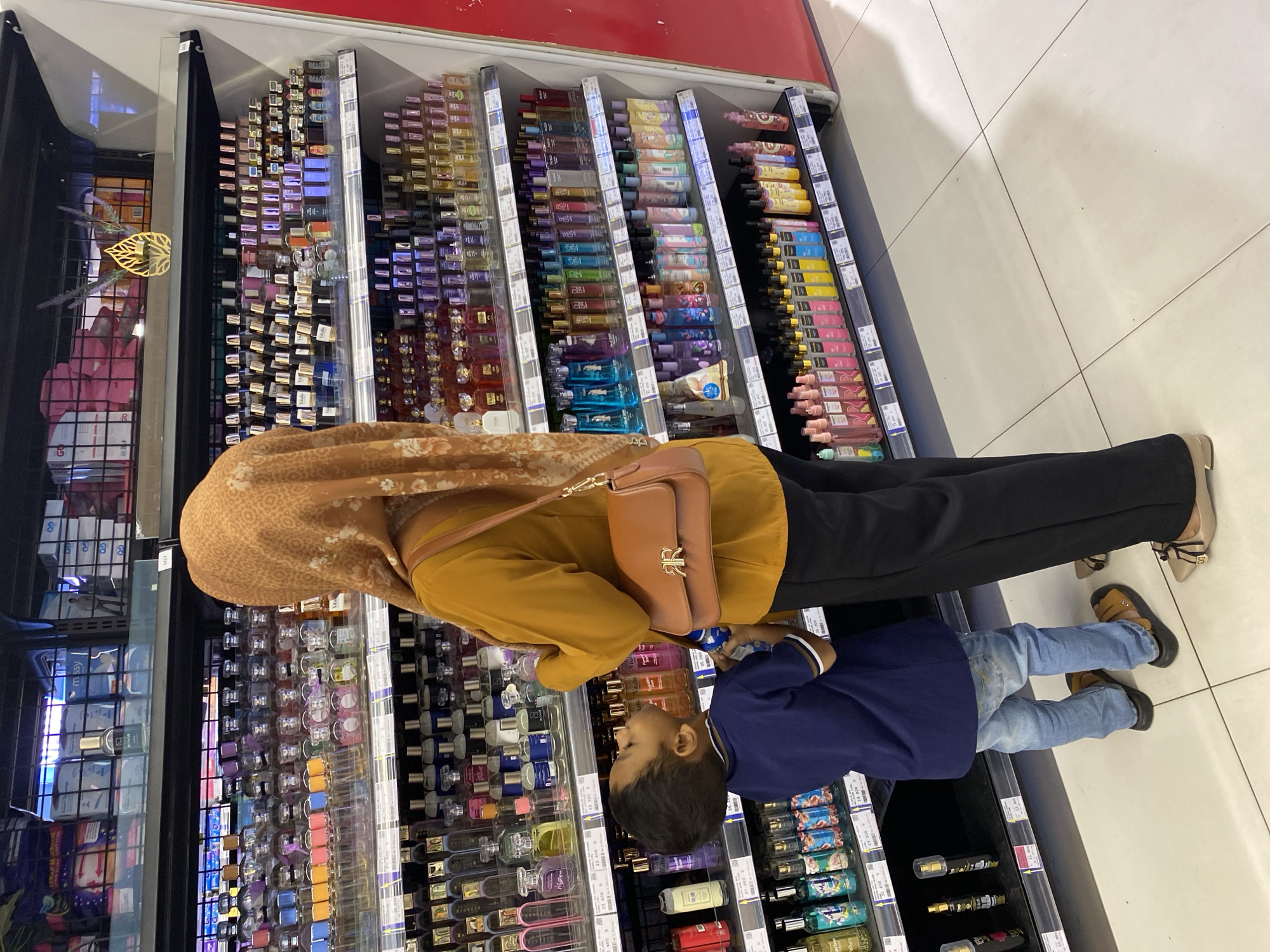 An adult wearing a mustard-colored long-sleeved shirt, black pants, and a patterned orange hijab is standing in a store aisle, looking at a display of perfumes. A young child in a blue shirt and jeans is standing next to the adult, looking up at the same display. Both are facing away from the camera. The aisle is lined with shelves filled with various bottles of perfume, cologne, and body sprays. The store appears to be a retail establishment in Banda Aceh, Indonesia, indicated by the price tags visible in Indonesian Rupiah on the shelves. The lighting is bright, typical of a retail environment.