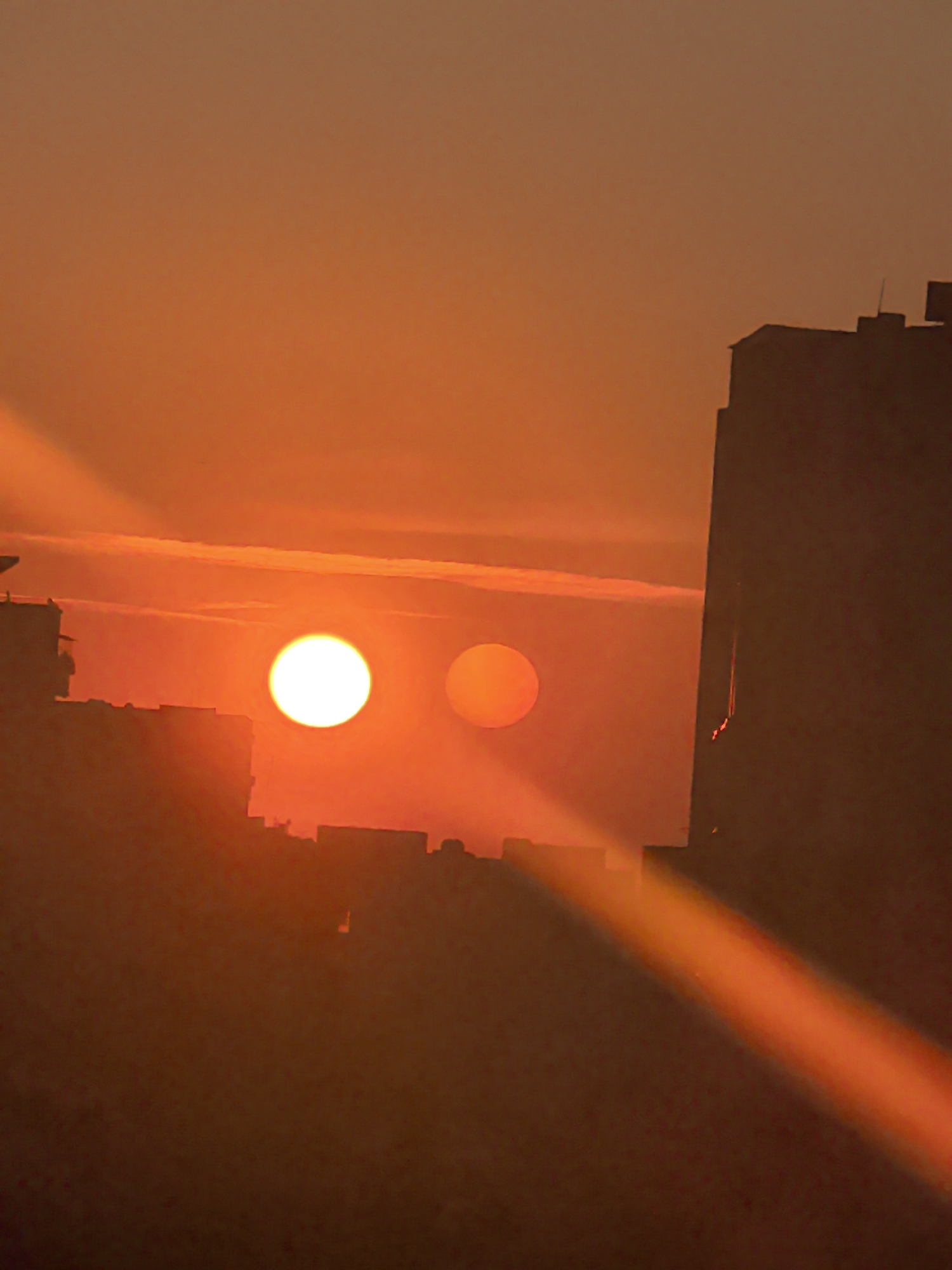 The image captures a vibrant orange sunset in what is likely Stara Zagora, Bulgaria, given the request. The sun is visible as a bright white orb, while a second, less distinct, orange-hued orb sits beside it. These are positioned behind a dark silhouette of buildings, typical of a city skyline. In the foreground, there is a blurred silhouette of an object, possibly a building, creating a sense of depth. A prominent beam of light shoots diagonally across the bottom portion of the image, further emphasizing the strong sunset colors and possibly highlighting atmospheric particles.
