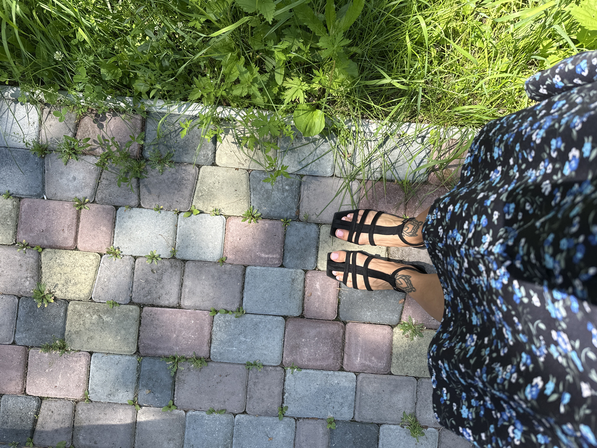 A high-angle shot shows a person's feet and legs wearing black sandals and a floral dress, standing on a multi-colored brick pathway next to a patch of vibrant green grass. The pathway is made of rectangular bricks in shades of gray, pink, and yellow, with small weeds growing in the cracks. The grass on the right is lush and appears healthy, with some small white flowers visible. The scene appears to be outdoors, likely in a garden or park setting. The location is identified as Duvankul', Russia, though there are no specific landmarks to confirm this from the image alone.
