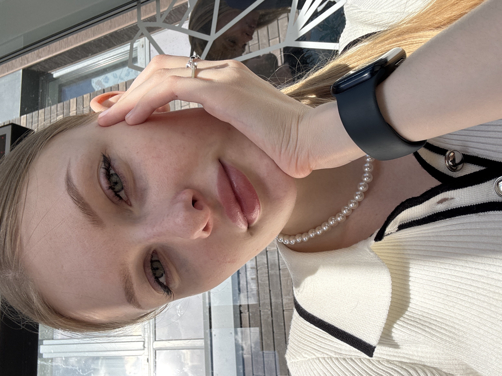 A young woman with blonde hair and green eyes is taking a selfie outdoors. She is wearing a white ribbed sweater with black trim, a pearl necklace, a smart watch on her left wrist, and a ring on her right ring finger. Her right hand is resting on her cheek, and her left hand is partially visible near her ear. The background shows a brick wall and some outdoor seating with a geometric metal design. The lighting suggests it is daytime. Based on the context provided of Perm, Russia, this could be an outdoor cafe or public space in the city.