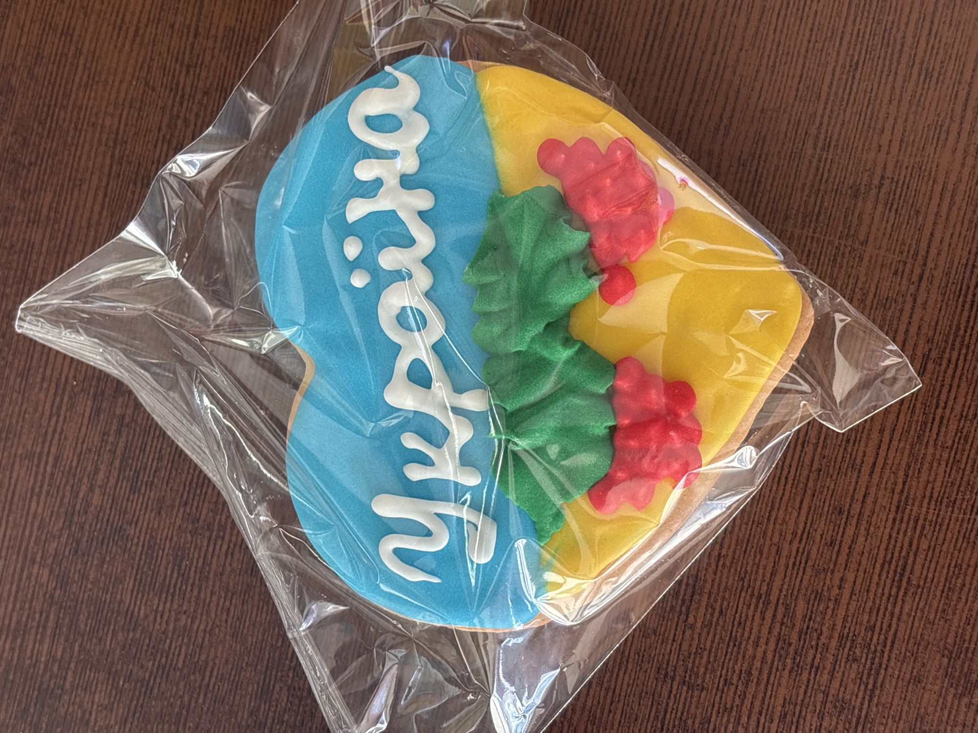 An overhead shot captures a heart-shaped cookie decorated with the Ukrainian flag colors and the word "Україна" in white icing. The cookie is enclosed in clear plastic packaging and rests on a dark, wooden surface. The top portion of the cookie is a vibrant blue, representing the sky, while the bottom is a bright yellow, symbolizing wheat fields. In the center, green fondant leaves and red berries are arranged, adding a decorative touch. The overall presentation is simple and patriotic.