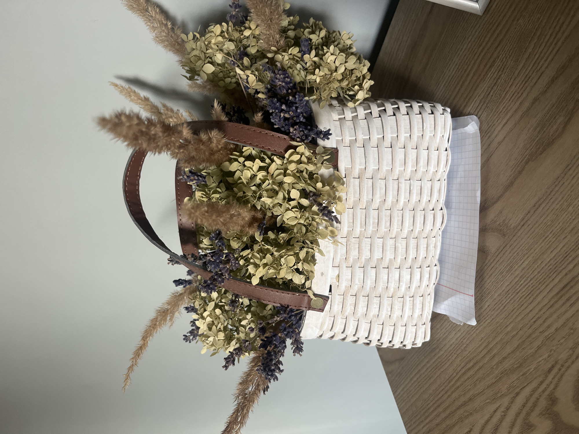 A close-up, eye-level shot shows a woven white basket filled with dried flowers and grasses. The basket has a brown leather handle. The arrangement is placed on a wooden surface, possibly a desk or table. Underneath the basket, a piece of graph paper is visible. The background is a plain, light-colored wall. The scene is indoors. The image does not depict any specific location in the city of Столбище, Russia, nor does it contain any people or actions.