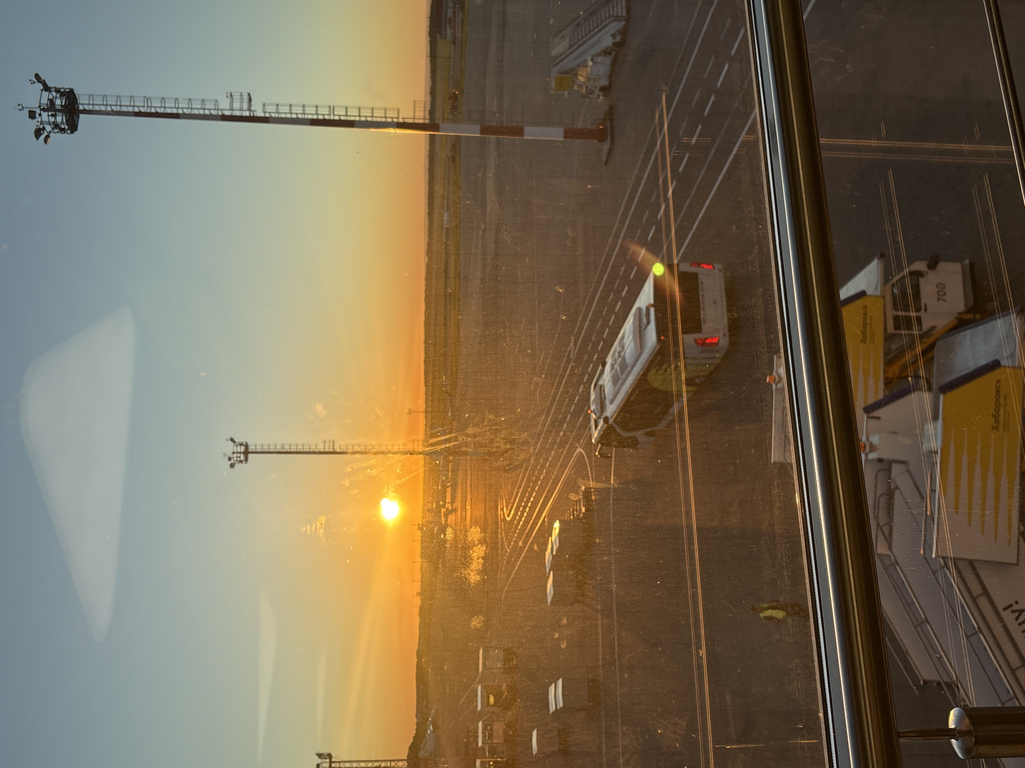 The image depicts an airport tarmac at sunset, with the sun low on the horizon and casting a warm golden glow across the scene. The location is identified as Khabarovsk, Russia, through text visible on an airport vehicle in the foreground.
In the midground, an airport bus, likely for passenger transport, is visible on the tarmac. Several cargo or luggage carts are parked on the left side of the image. In the background, a series of airport lighting towers stand tall, their structures silhouetted against the vibrant sky. The scene is viewed through a window, which shows some smudges and reflections, characteristic of an indoor vantage point.
In the foreground, a metal railing is visible, suggesting the viewpoint is from an elevated area like an airport terminal building or observation deck. Part of an airport staircase or jet bridge is also present in the bottom section of the frame, with the number "700" visible on a vehicle beneath it. A person wearing a high-visibility vest can be seen standing on the tarmac near some parked carts. The overall environment is that of an active airport during the evening hours.