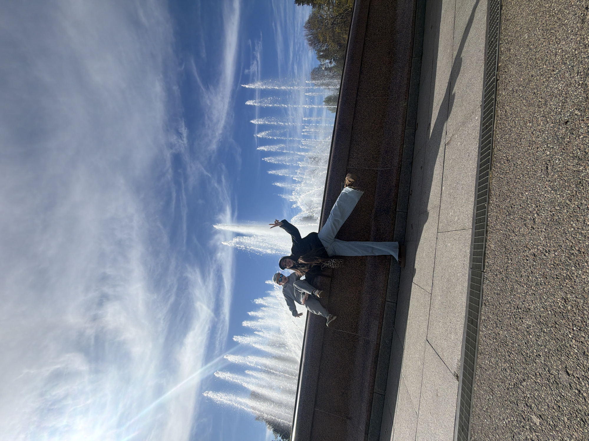 An outdoor scene captures two individuals posing on a stone ledge beside a series of active water fountains under a bright, sunny sky. The image is taken on a clear day, indicated by the vibrant blue sky adorned with wispy white clouds.
On the right side of the frame, two people are seated on a wide, dark stone wall or edge, possibly part of a large fountain complex. The person in the foreground wears light blue pants, a dark jacket with a leopard print detail, and a dark hat, striking a playful pose with one leg extended upwards and one arm forming a peace sign. Behind them, another individual is partially visible, wearing a light-colored hoodie and sunglasses, also appearing to pose.
Behind the people, a grand fountain display features numerous individual water jets spraying high into the air, creating a dynamic backdrop. Beyond the fountains, some trees with bare branches are visible against the sky, suggesting an early spring or late autumn setting.
The ground area to the right of the stone wall is paved with light grey tiles, adjacent to a long metal grate, and further right, a darker, granular pavement. Long shadows stretch across the ground, indicating the sun is relatively low in the sky, likely mid-morning or late afternoon. The overall impression is one of leisure and enjoyment in a public urban setting, potentially a park or square in Saint Petersburg, Russia. No visible text is present in the image.