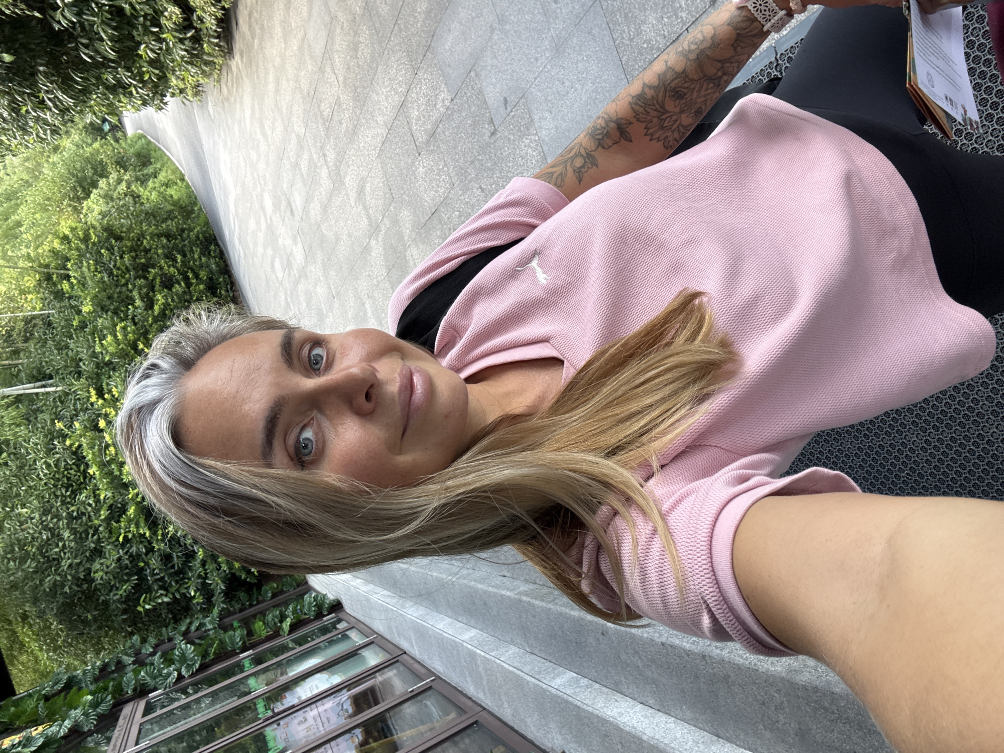 A blonde woman with grey streaks in her hair, blue eyes, and a light complexion is captured in a selfie. She is wearing a pink Puma t-shirt and dark leggings. Her right arm, visible on the right side of the frame, features floral tattoos. She is seated outdoors on a patterned bench with a black bag on her shoulder. In the background, a paved walkway curves to the right, flanked by lush green trees and foliage. To the left of the woman, a building with large windows covered in green plants is visible. The overall scene suggests an outdoor setting within a city park or a similar urban green space in Wuxing District, China.