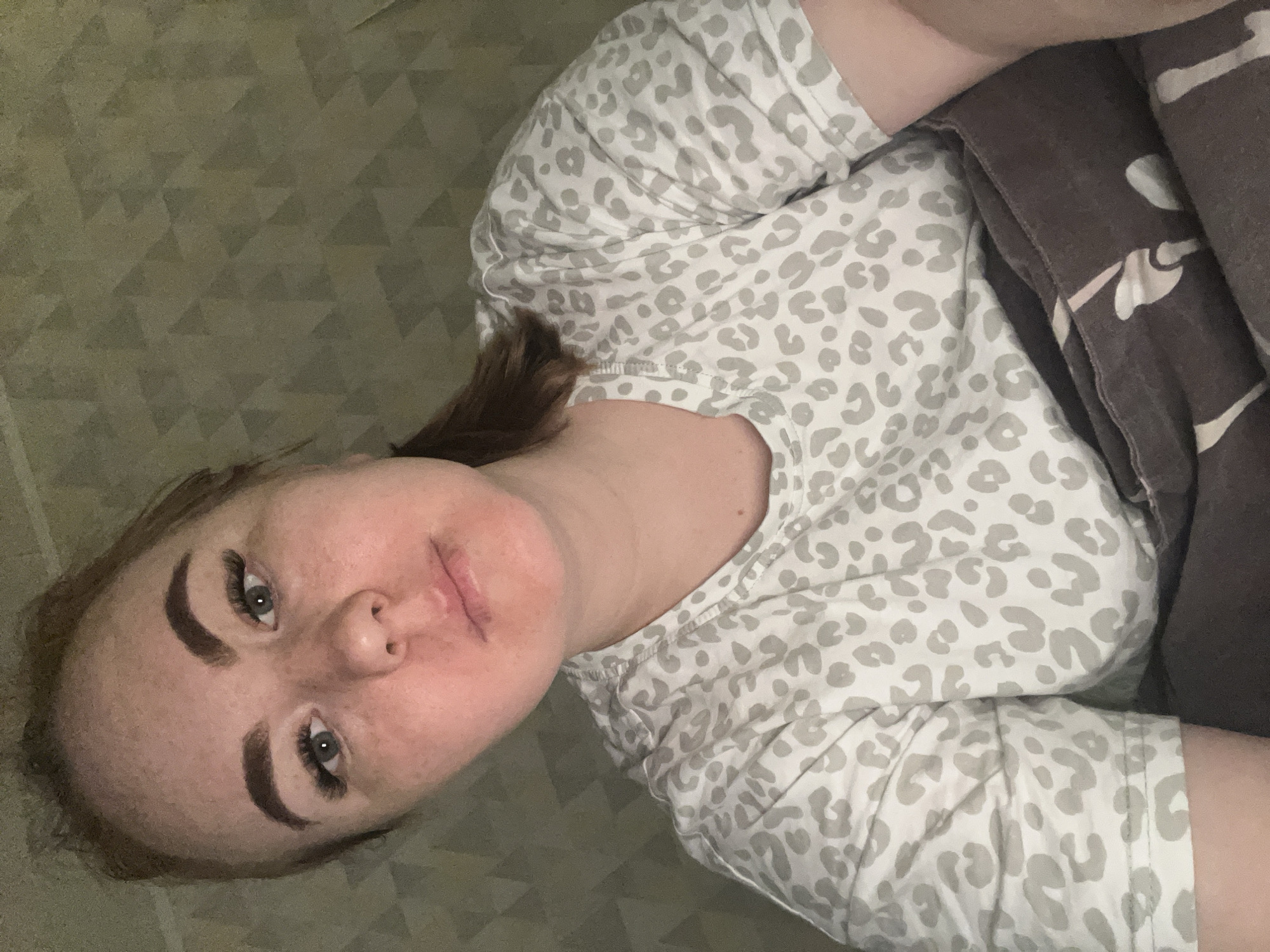 A woman is lying down, possibly on a bed or sofa, covered with a brown and white blanket. She is wearing a light gray and white animal print shirt. The background includes a patterned surface, likely the bedspread or floor. There's no contextual information that would place this image in Chaikovsky, Russia.