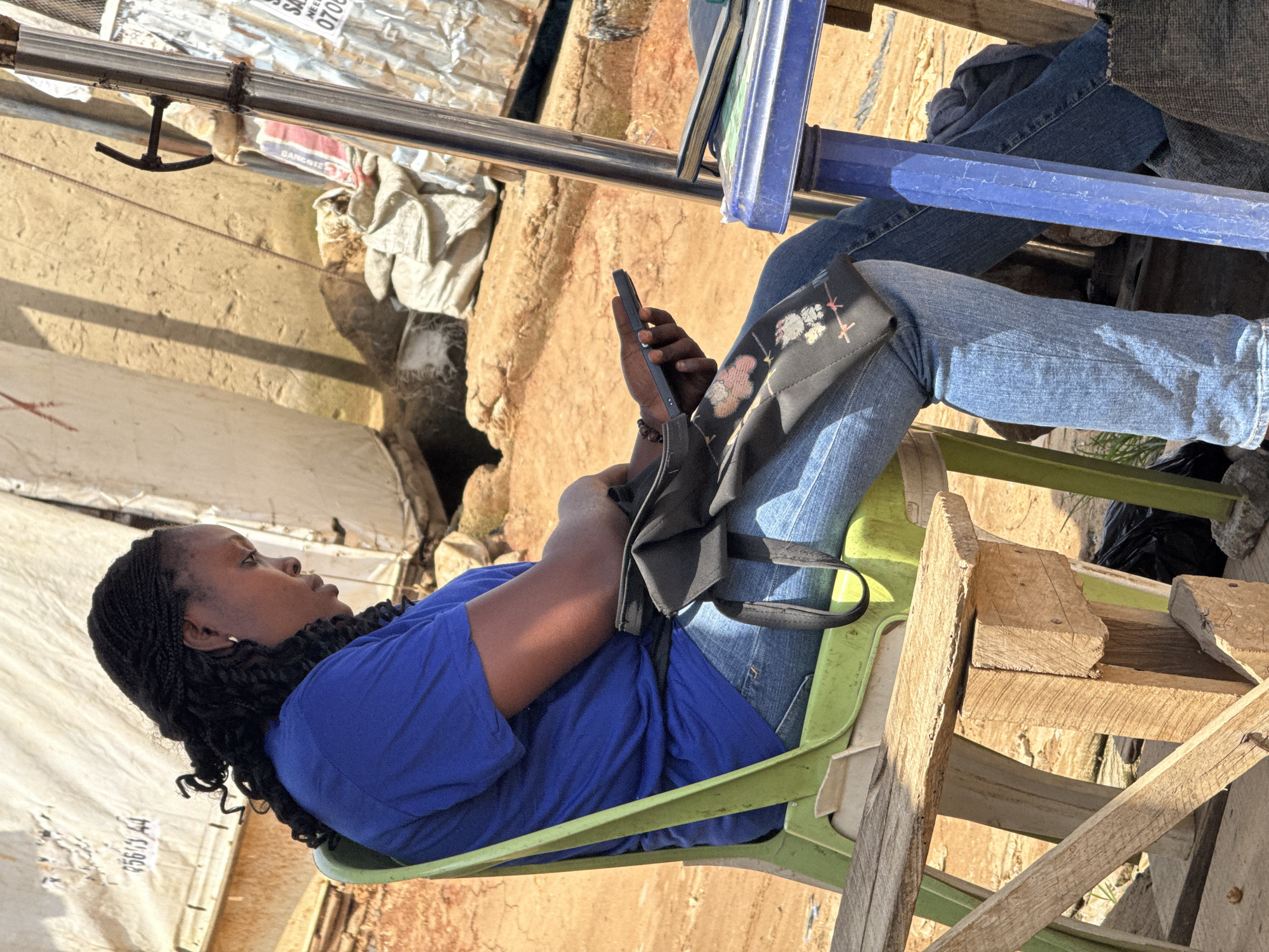 In Abuja, Nigeria, a woman with braided hair is seated on a light green plastic chair. She is wearing a blue t-shirt and blue denim jeans. A black bag with a floral design is draped over her lap, and she is holding a smartphone in her right hand. The woman is looking down at the phone, seemingly engaged with it. She is seated next to a blue table, on which a closed book or notebook is placed. The background of the image shows a rough, outdoor environment with sandy ground, some fabric structures, and a metal pole. Sunlight casts shadows on the scene, indicating it is daytime.