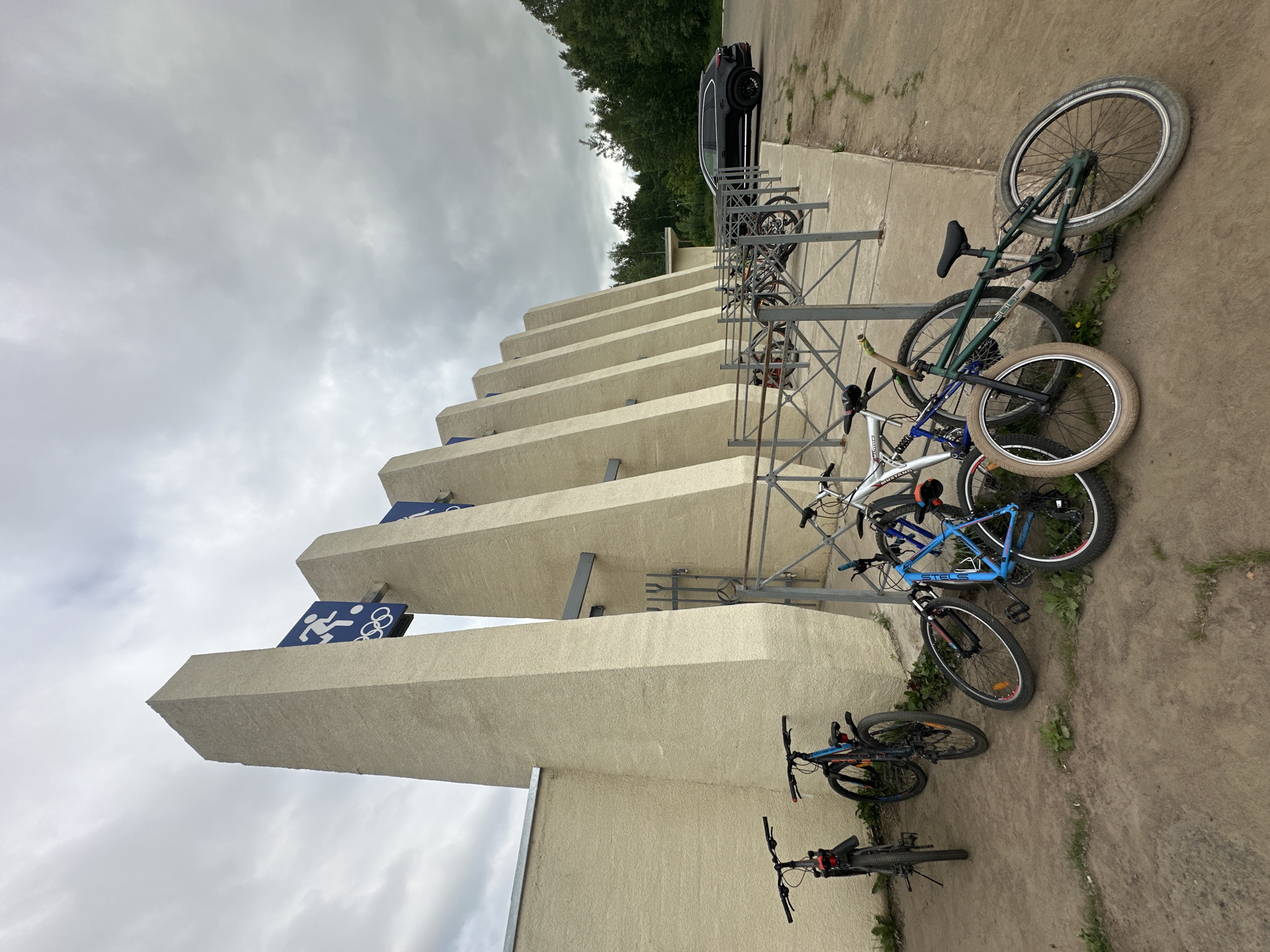 The image shows a bicycle parking area outside a building with a series of concrete pillars. The pillars are tall and angular, with blue signs displaying the Olympic rings and silhouettes of athletes engaged in sports. Several bicycles are parked in front of the building. Two black bicycles are parked on the left, one with a red reflector on the rear. Further to the right, a blue bicycle is leaning against a metal fence, and a white bicycle is also visible. Another green bicycle is parked on the ground in the foreground. A black SUV is parked to the right of the building, and a treeline is visible in the background. The sky is overcast. This scene is located at the Chaikovsky Sports Arena in Chaikovsky, Russia, a facility associated with the 1972 Winter Olympics.