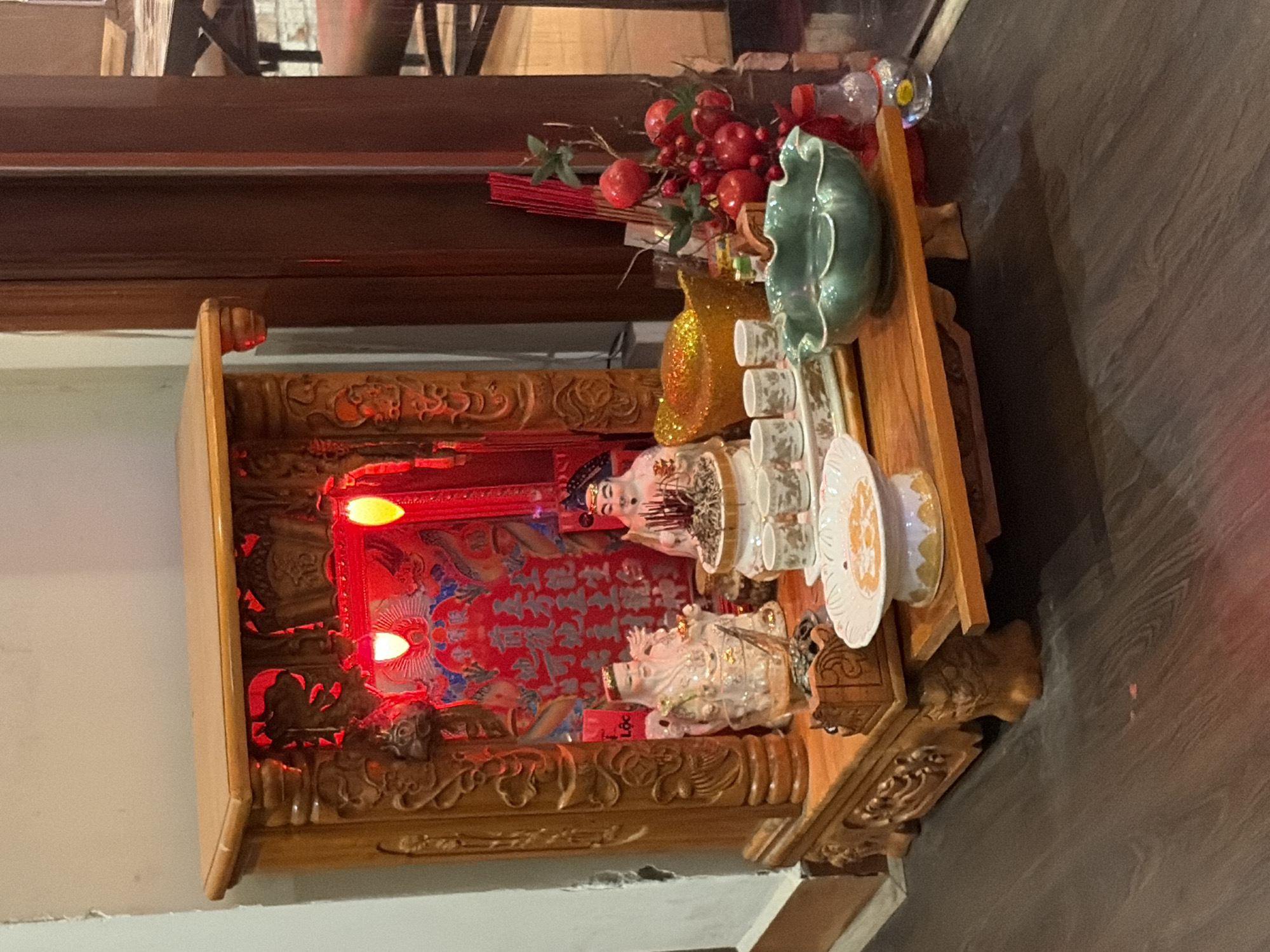 A wooden altar is positioned on a dark wooden floor in Thủ Đức City, Vietnam. The altar is ornately carved and illuminated by red lights from within. On the altar, there are two ceramic figurines, one white and one pink, resembling deities or revered figures. In front of them, a gold-colored object, possibly a wealth symbol, is placed. A collection of small white cups and saucers sits to the right of the figurines, along with a bowl filled with what appears to be dried herbs or incense. A cluster of red artificial fruits on branches and a bunch of red incense sticks are also present. A green, ruffled bowl is situated in front of the incense sticks, and a small glass bottle with a red cap is to its right. A white plate with a decorative pattern is placed at the lower right of the altar. The background features a portion of a dark wooden door or partition and a glimpse of a dining area with furniture.