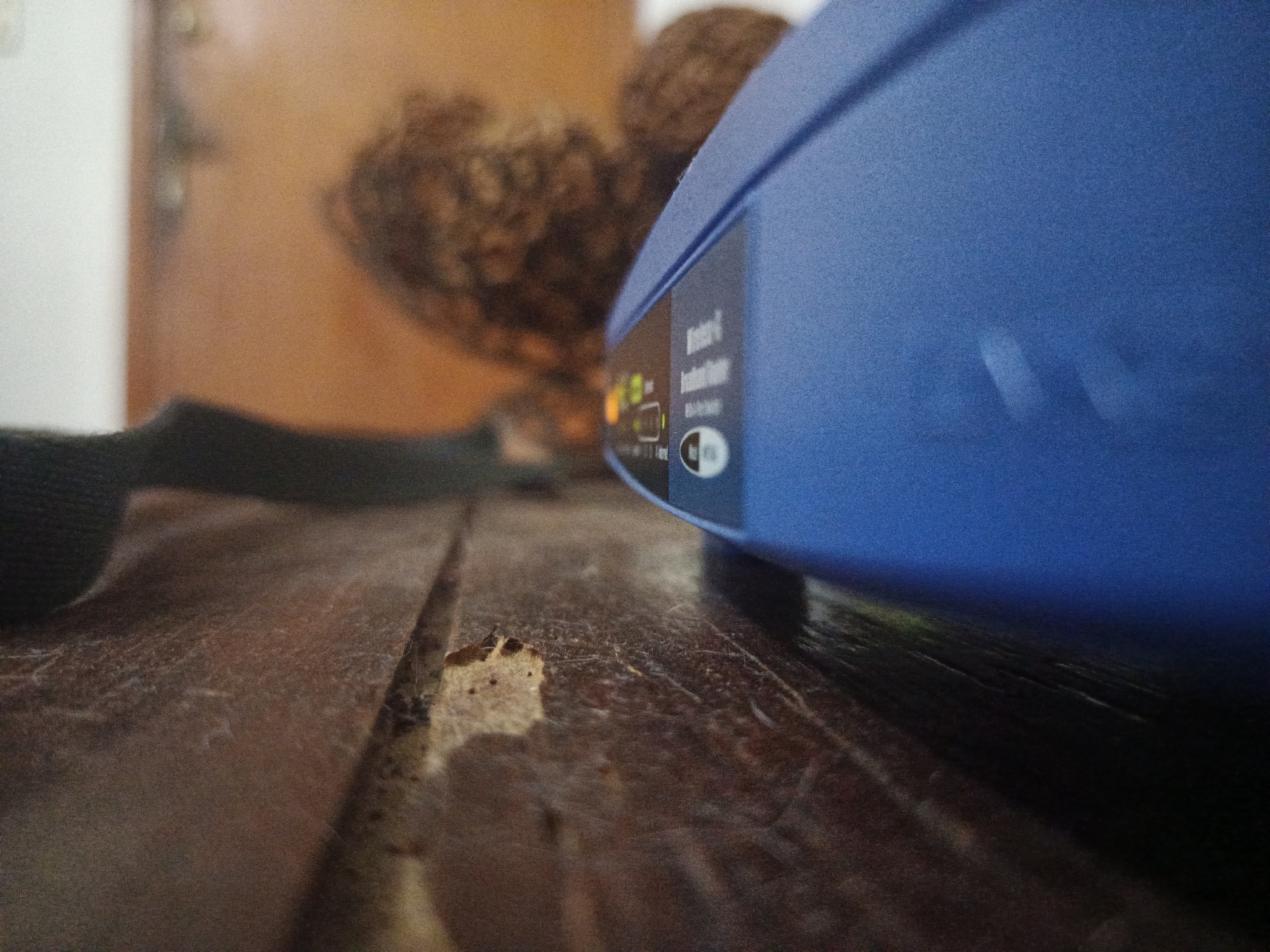 A close-up shot focuses on a dark wooden surface with a prominent crack and loose debris in the foreground. To the right, a blue electronic device, possibly a router, sits partially in the frame, displaying illuminated indicator lights and printed text. In the background, a blurry brown object and a light-colored wooden door or wall with a visible handle add depth to the scene. A black strap is partially visible on the left side of the frame. This scene cannot be located in Los Robles, Venezuela, based on the available visual information.