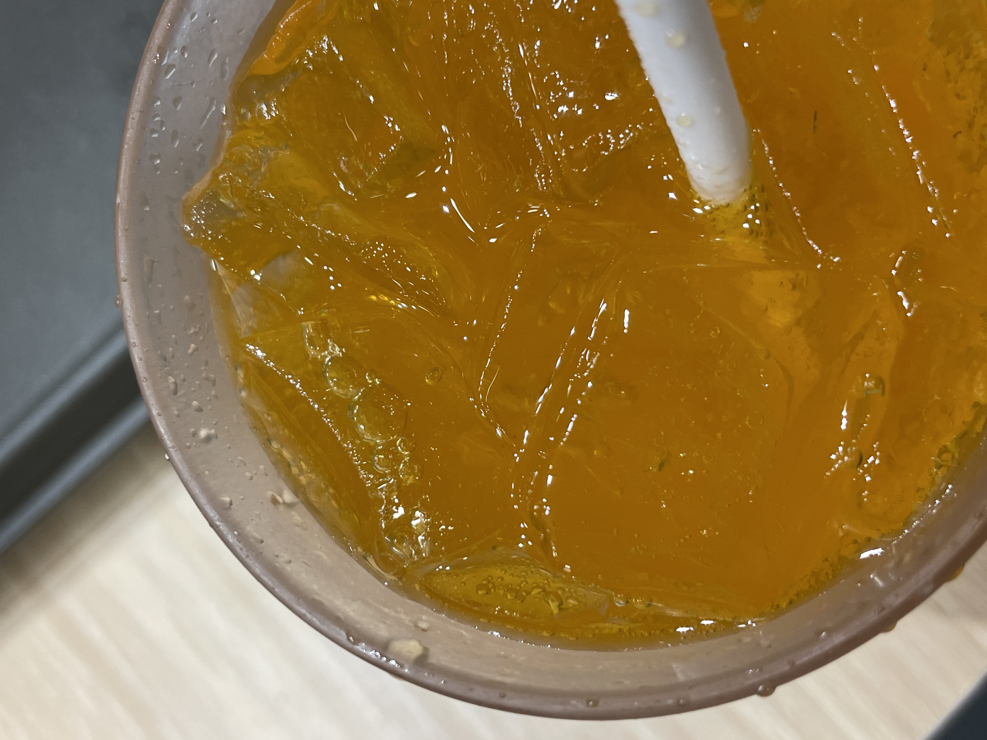 The image provides a close-up, top-down view of a cold beverage served in a translucent plastic cup. The cup's exterior is covered in small condensation droplets, indicating the drink is chilled. Inside, a vibrant orange-colored liquid is filled with numerous clear ice cubes that sparkle as they reflect ambient light. A white plastic straw is inserted into the drink, angled from the top right, with its lower end submerged. Small bubbles are visible around the base of the straw where it enters the liquid. The background is out of focus, showing blurred light-colored surfaces, possibly a table or counter, and darker, indistinct elements, suggesting an indoor setting. There are no people visible, nor is there any discernible text within the frame. The image captures a refreshing drink ready for consumption.