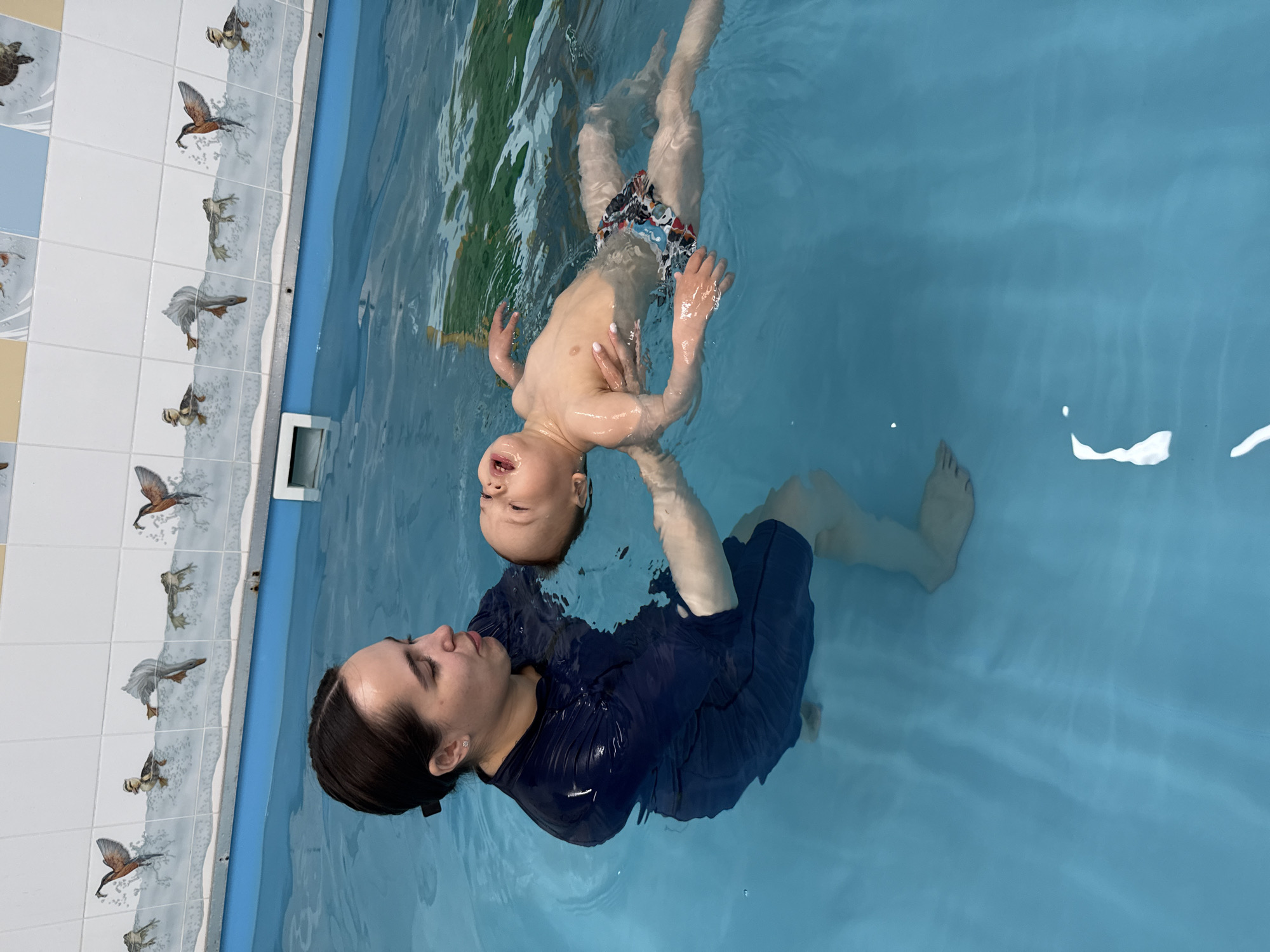 An overhead shot captures an adult and a young child in a light blue swimming pool. The adult, a female with dark hair pulled back and wearing a dark blue long-sleeved garment, is submerged to her shoulders, attentively looking towards the child. She is supporting the child from beneath, with her right hand under the child's back and her left arm gently around their side.
The child, a toddler wearing patterned swimming trunks, is floating on their back, seemingly held by the adult. Their arms are slightly raised, and their mouth is open as if in mid-expression, perhaps vocalizing or reacting to the water. The child's bare torso and legs are visible through the clear blue water, which shows gentle ripples.
The edge of the pool is visible on the left side of the frame, featuring distinctive white tiles decorated with illustrations of various animals, including ducks in flight, frogs, and fish, some seemingly in motion with splashes. Beneath these illustrations, there is a patterned border that mimics water or foam. Below the white tiles, there is a section of solid light blue tiling, and further down, some light yellow/tan tiles. A white rectangular grate or skimmer is visible on the pool wall. The scene suggests a swimming lesson or playful interaction in a pool setting during daylight hours. No visible text is present.