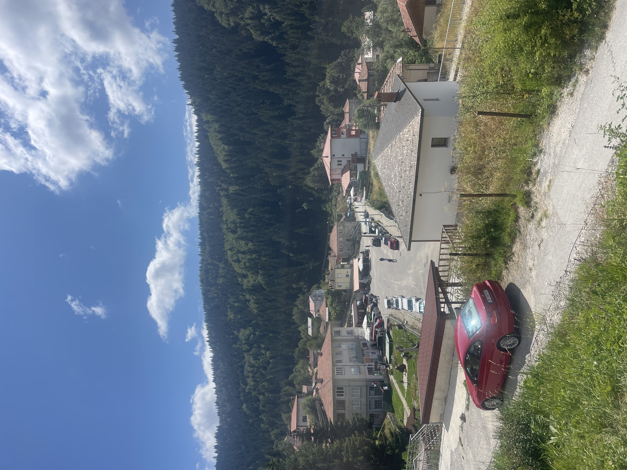 The image shows a village nestled in a mountainous region, possibly in the Rhodope Mountains near Manastir, Bulgaria. The foreground features a red BMW coupe parked on a dirt road, surrounded by lush green grass and vegetation. A gravel pathway leads up to the car. Beyond the car, a paved road winds through the village, lined with several parked cars. Buildings with red-tiled roofs are visible, some appearing to be residential homes and others possibly small businesses. The architecture suggests a traditional Balkan style. In the background, a dense forest of pine trees covers a steep mountainside, reaching up to a bright blue sky dotted with white clouds. The overall scene depicts a peaceful, rural setting in a natural, mountainous landscape.