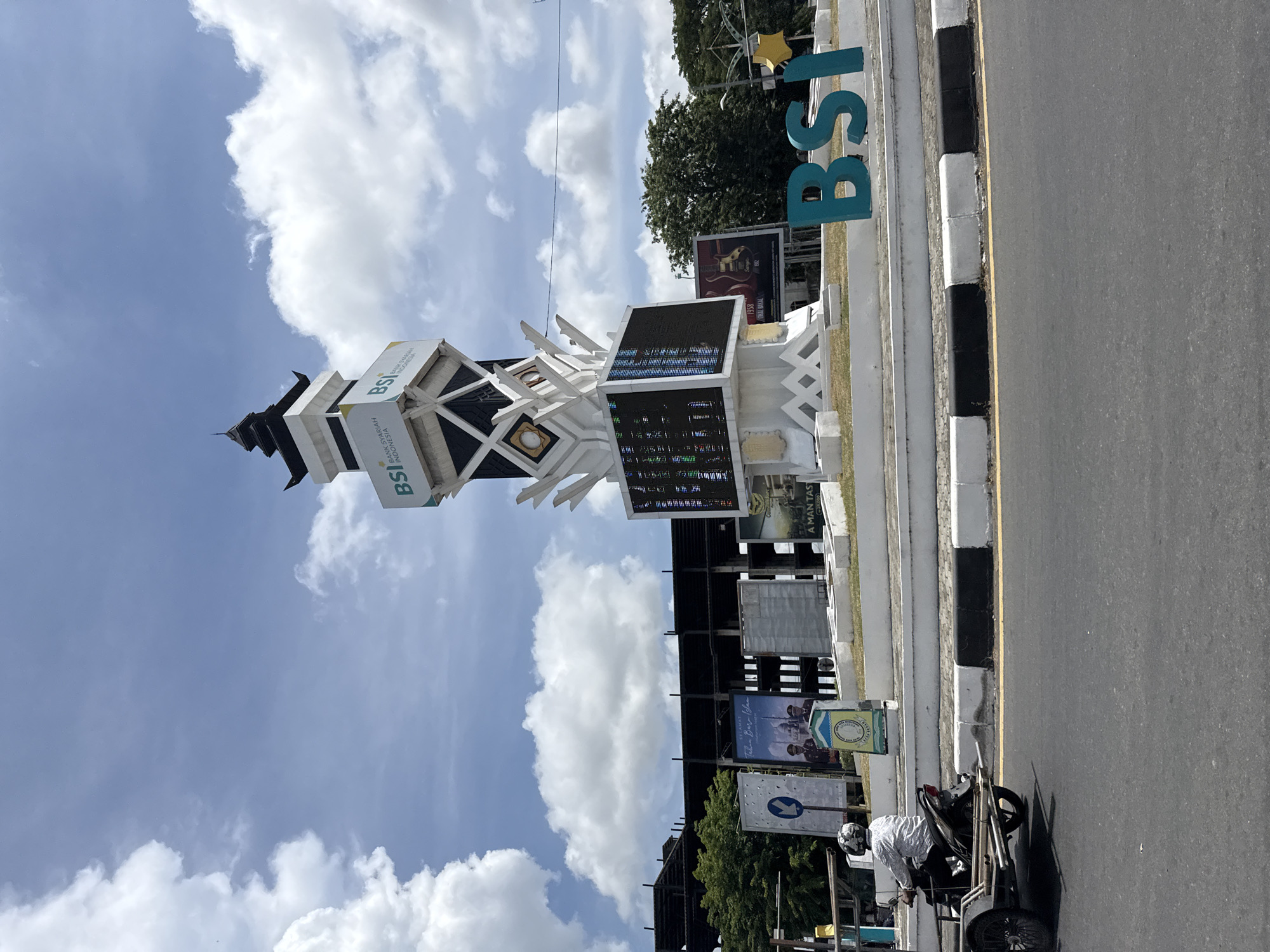 In Lambaro, Indonesia, a modern monument with the "BSI" logo stands tall against a partly cloudy sky. The monument features multiple tiers, with the "BSI" branding prominently displayed on its upper sections. A large digital screen is integrated into the monument's base, showcasing a complex array of colorful pixels, suggesting it may be displaying data or advertisements. To the right of the monument, large, teal-colored "BSI" letters are positioned on the ground, further reinforcing the branding. In the background, a partially constructed building, likely a commercial or public structure, is visible, with its skeletal framework exposed. Several billboards are also present, one featuring an advertisement with two figures in uniform and text that appears to read "SELAMAT TAHUN BARU." To the left of the monument, a motorcycle with a small trailer attached is moving away from the camera, driven by a person wearing a helmet and a patterned shirt. The road in the foreground is asphalt, marked with a yellow line and a black and white striped curb. Lush green trees are visible behind the billboards and the construction site, indicating a well-developed urban environment. The overall scene depicts a busy public space with a focus on commercial branding and urban development.