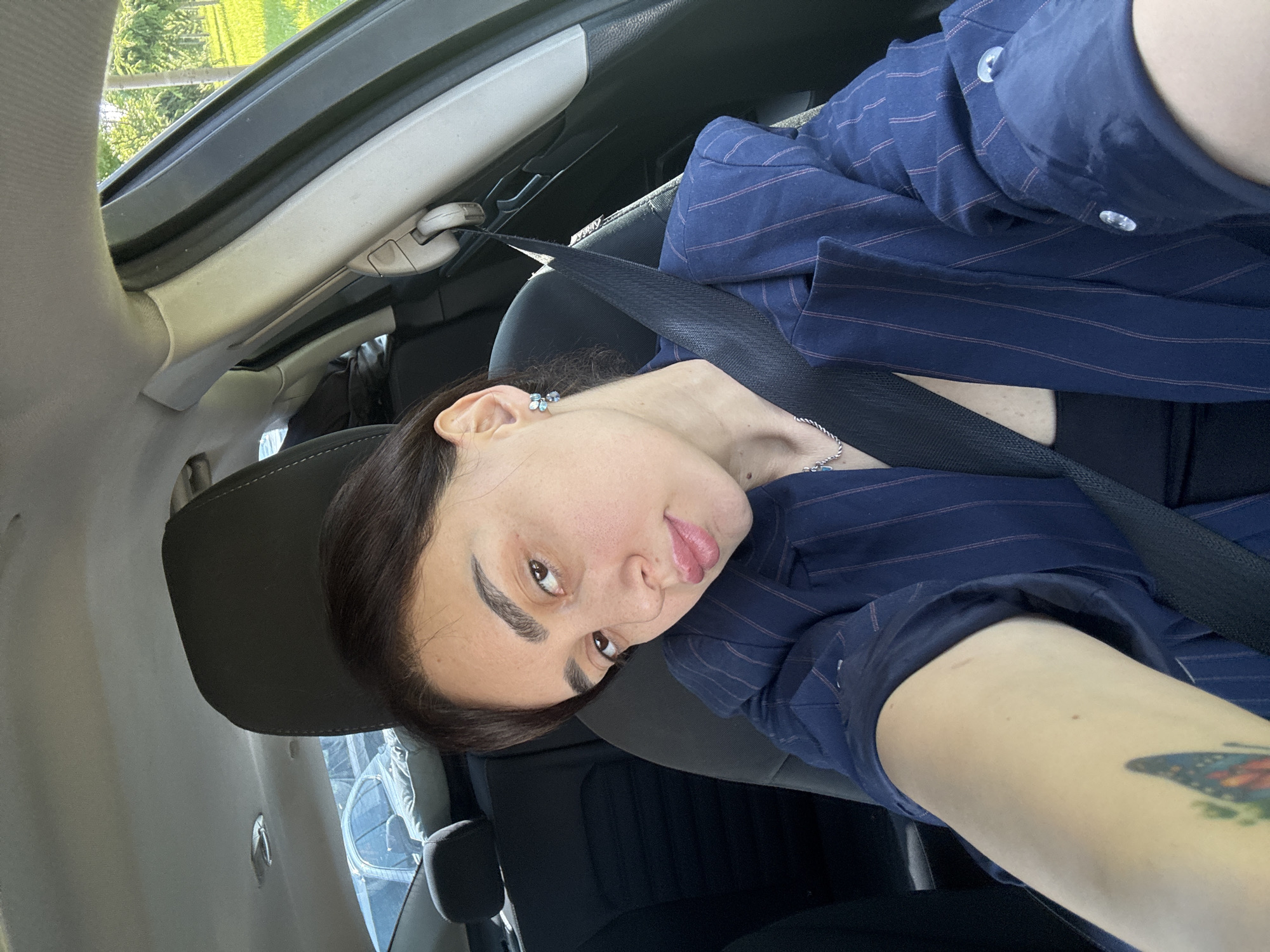 A selfie image shows a person in a car. The person is wearing a blue pinstripe shirt and a seatbelt. The person has dark hair and fair skin. Part of a tattoo is visible on their left forearm. The interior of the car is visible, including the dashboard and seats. Outside the car, green foliage is seen, blurred, possibly trees. The location is undetermined.
