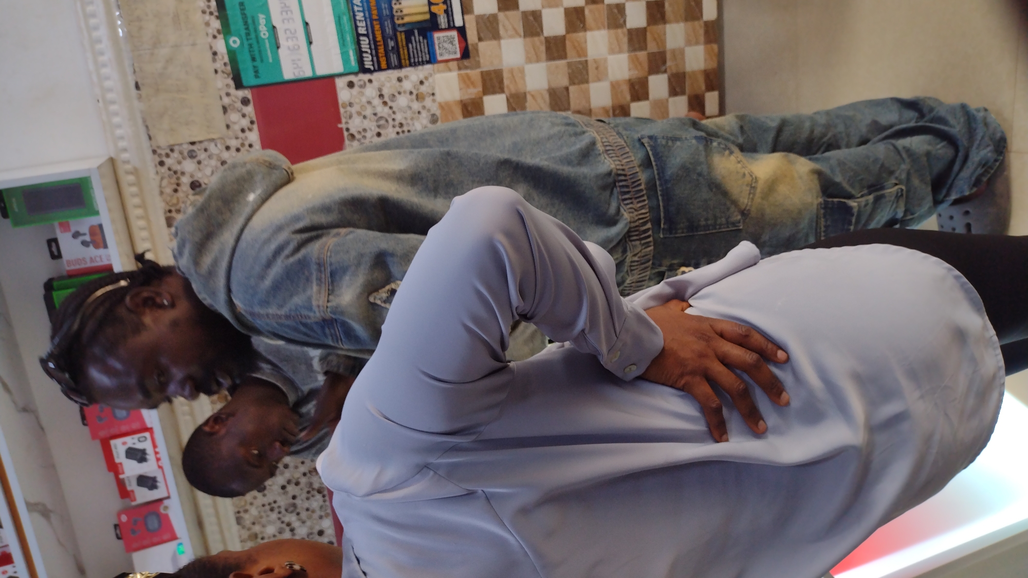 Two dark-skinned men are in a room in Jos, Nigeria. One man has braids and is wearing a denim jacket and jeans. The other man is wearing a light blue shirt. They are talking. The room has tiled walls and a display of electronic devices. There is a poster for "JIUJIU RENTAL" on the wall.