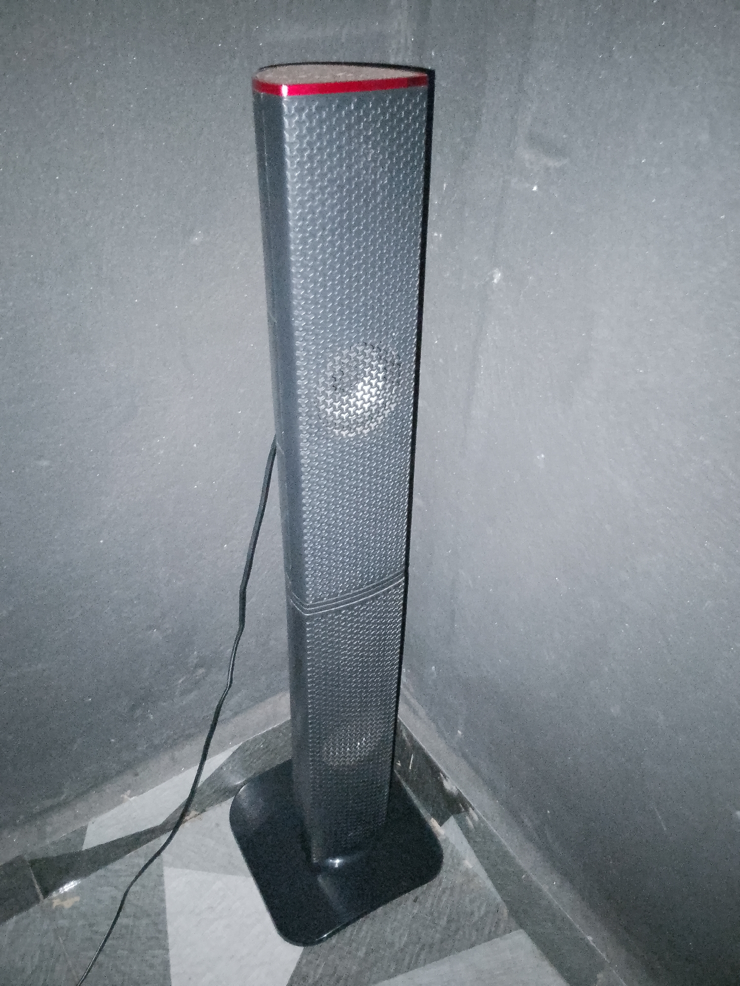 A tall, narrow, dark gray speaker stands upright in the corner of a room. The speaker has a textured surface with a pattern of small raised dots and a red band across its top. A black wire extends from the speaker and runs along the floor. The floor has a diamond pattern of dark gray and light gray tiles. The walls appear to be dark gray and textured. The scene is dimly lit.
