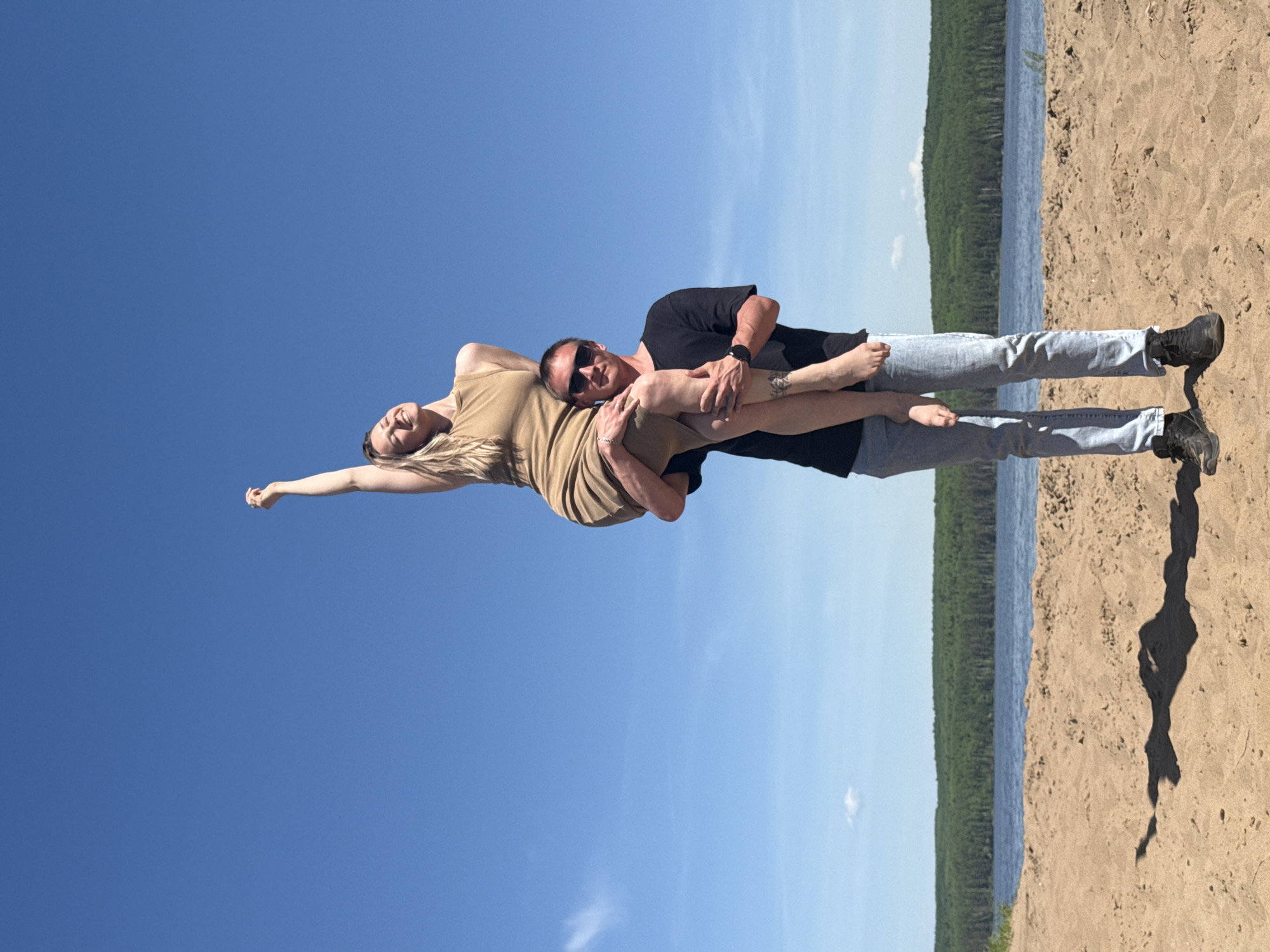 A man and a woman are posing for a photo on a beach. The man, wearing a black t-shirt, light blue jeans, and dark shoes, is holding the woman in his arms. She is wearing a tan dress. The man is also wearing sunglasses and a wristwatch. The background shows a body of water with a forested shoreline under a clear blue sky. The location is near the city of Chaikovsky, Russia. The man is standing on the sand with his shadow cast beside him. The woman has one arm extended upwards.
