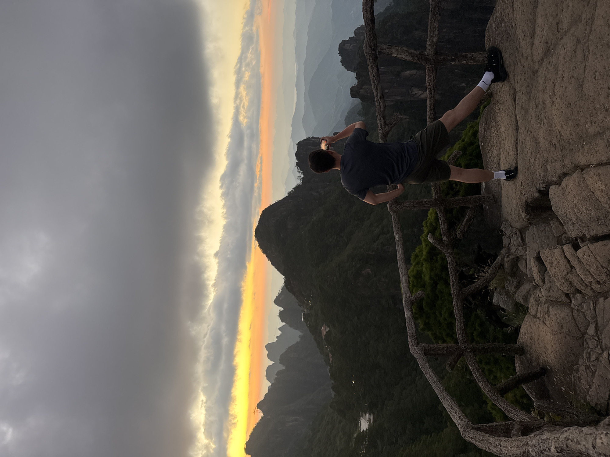 A person is standing on a rocky overlook, facing away from the camera, and taking a photo with their phone. They are wearing a dark t-shirt, olive green shorts, white socks, and black shoes. The person is positioned on the right side of the frame. In front of the person is a rustic wooden railing. The background features a dramatic mountain landscape during sunset. Jagged, dark green mountains stretch into the distance, with layers of misty, blue-tinged mountains fading towards the horizon. The sky is filled with dramatic, dark gray clouds, with a vibrant strip of orange and yellow light from the setting sun illuminating the horizon. The scene is likely located in Huangshan District, China, known for its iconic mountain scenery.