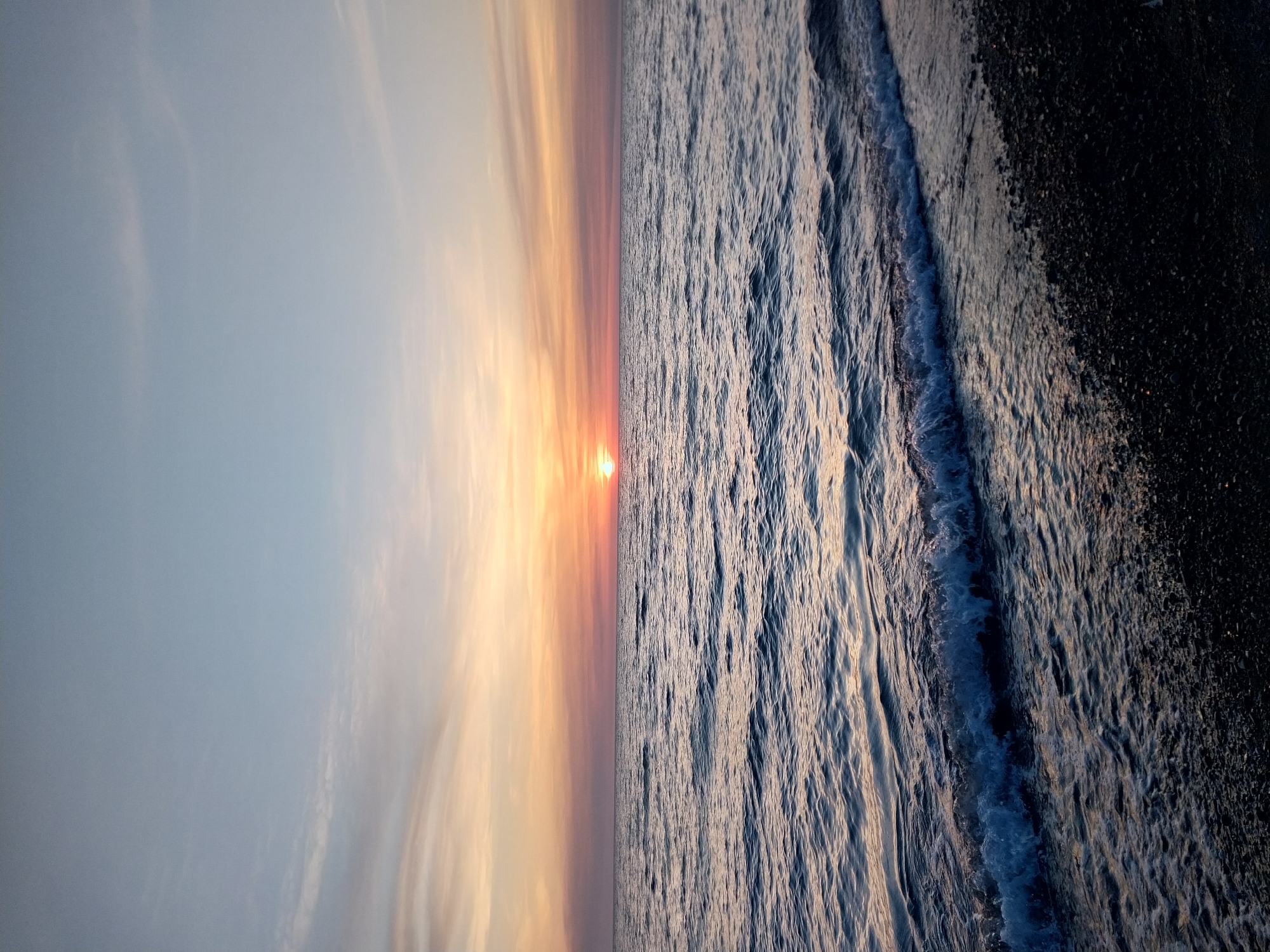 The image depicts a sunset over the Black Sea in Sochi, Russia. The sky on the left shows a gradient of light blue to orange, with soft, wispy clouds. The sun is visible on the horizon as a small, bright orange orb. The water on the right is textured with waves and ripples, transitioning from lighter, reflective areas to darker, shadowed parts. Close to the right edge, a strip of dark, pebbled beach is visible where the waves are breaking.