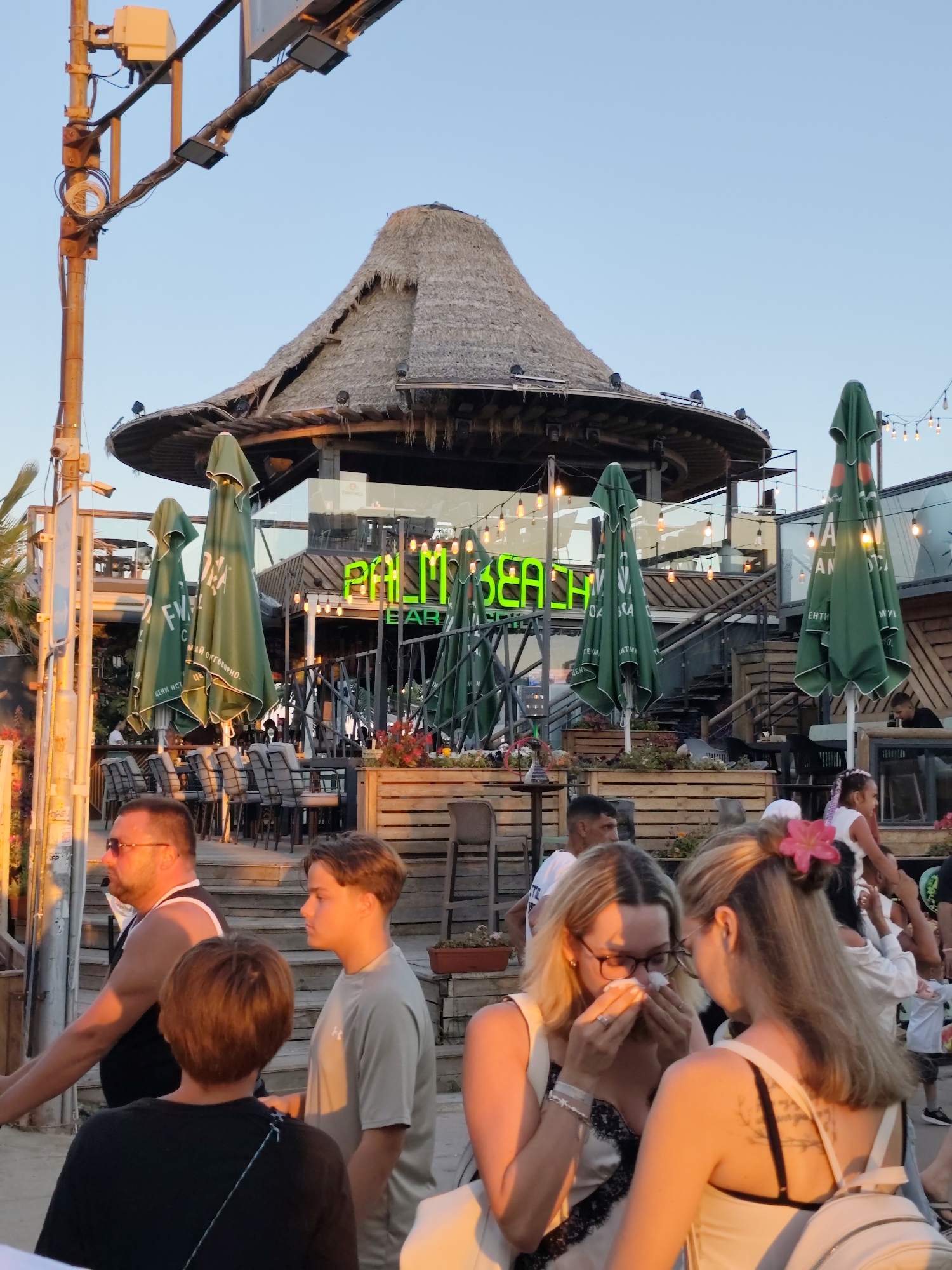 A group of people are gathered outside a beachfront establishment with a thatched roof in Nesebar, Bulgaria. In the foreground, a man wearing sunglasses and a black tank top is visible on the left, facing away from the camera. Next to him, a young boy in a grey t-shirt stands looking to the right. Another child with short brown hair is partially visible in the lower left foreground, facing away. In the middle ground, two women are interacting. One woman, wearing glasses, is holding a tissue to her nose and eyes. The other woman, with blonde hair adorned with a pink flower, is looking at the first woman. Behind them, green beach umbrellas are set up, and tables and chairs are visible under a structure with a thatched roof. Neon green lettering spelling out "PALM BEACH BAR" is illuminated on the building. String lights are strung across the scene, and palm trees are present in the background. The lighting suggests it is late afternoon or early evening.