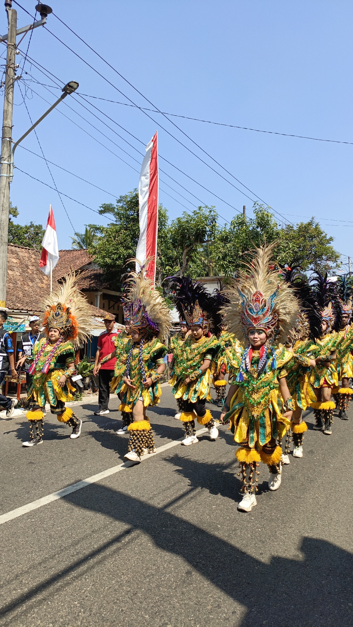 In Krajan Lor, Indonesia, a group of young people in elaborate, colorful costumes are participating in a parade. The costumes feature vibrant yellow, green, and blue fabrics, ornate headdresses with large feathers, and beaded leg coverings. They are walking on an asphalt road lined with trees and houses. Two large flags, one red and white and another white and red, are visible in the background, suggesting a national celebration. A utility pole with electrical wires and a street light stands to the left of the frame. People in the background are observing the parade. The scene is bathed in bright sunlight, casting long shadows on the road.