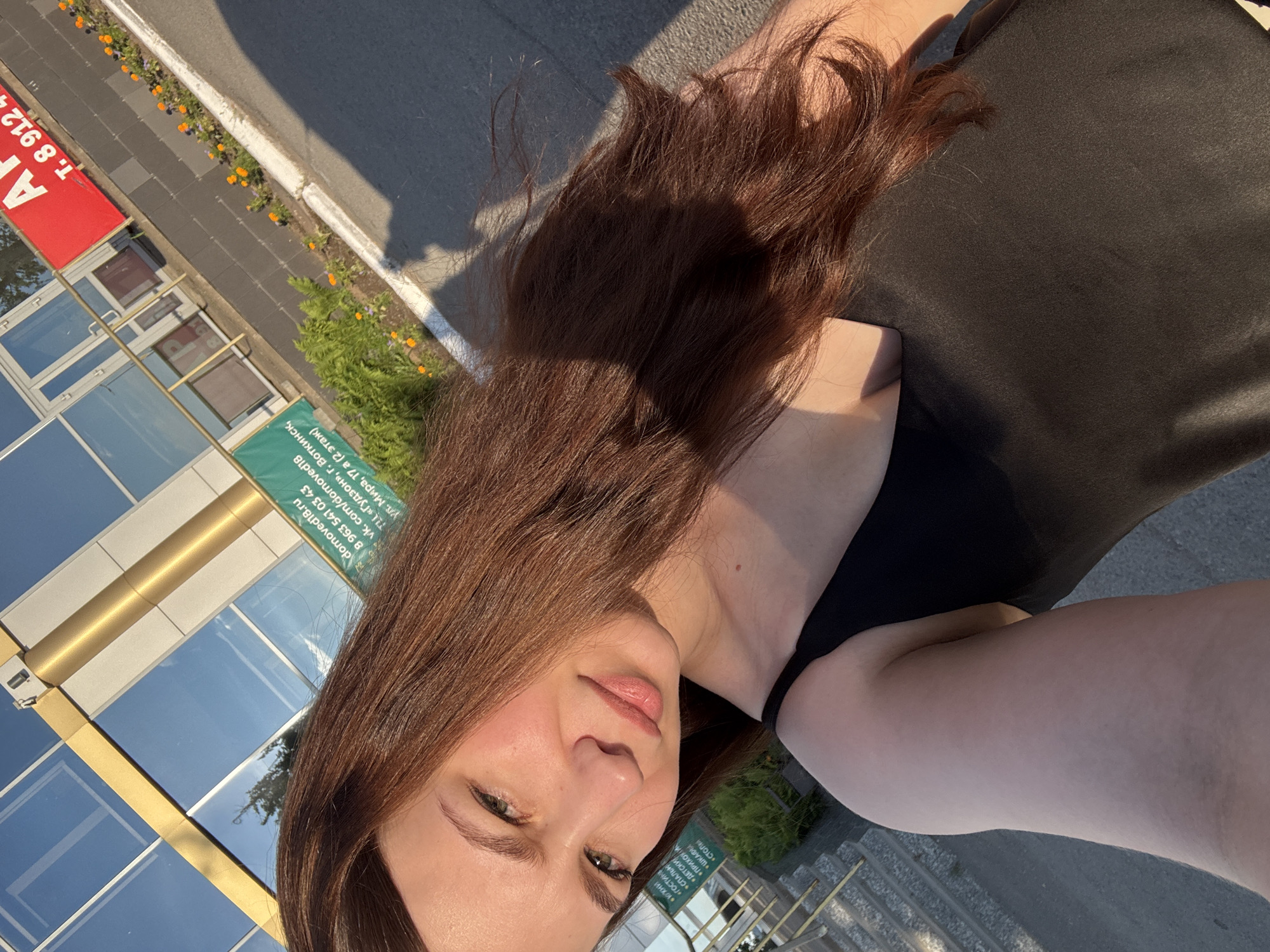 A young woman with long, dark auburn hair is taking a selfie outdoors in front of a building with a modern facade and reflective windows. She is wearing a black tank top. The building appears to be a commercial establishment, as evidenced by signage. One sign, partially visible in the image, displays a website address: "domoved18.ru", a phone number "8 963 541 03 43", and a VK.com handle "vk.com/domoved18". Another sign, in red and white, shows "AP" and a phone number that begins with "T. 8 912". Additionally, there is a green banner with text in Russian, mentioning "г. Воткинск" (city of Votkinsk). The ground in front of the building is paved, and there is a strip of landscaping with orange and purple flowers. The sun is casting a shadow of the woman onto the pavement, suggesting it is daytime.