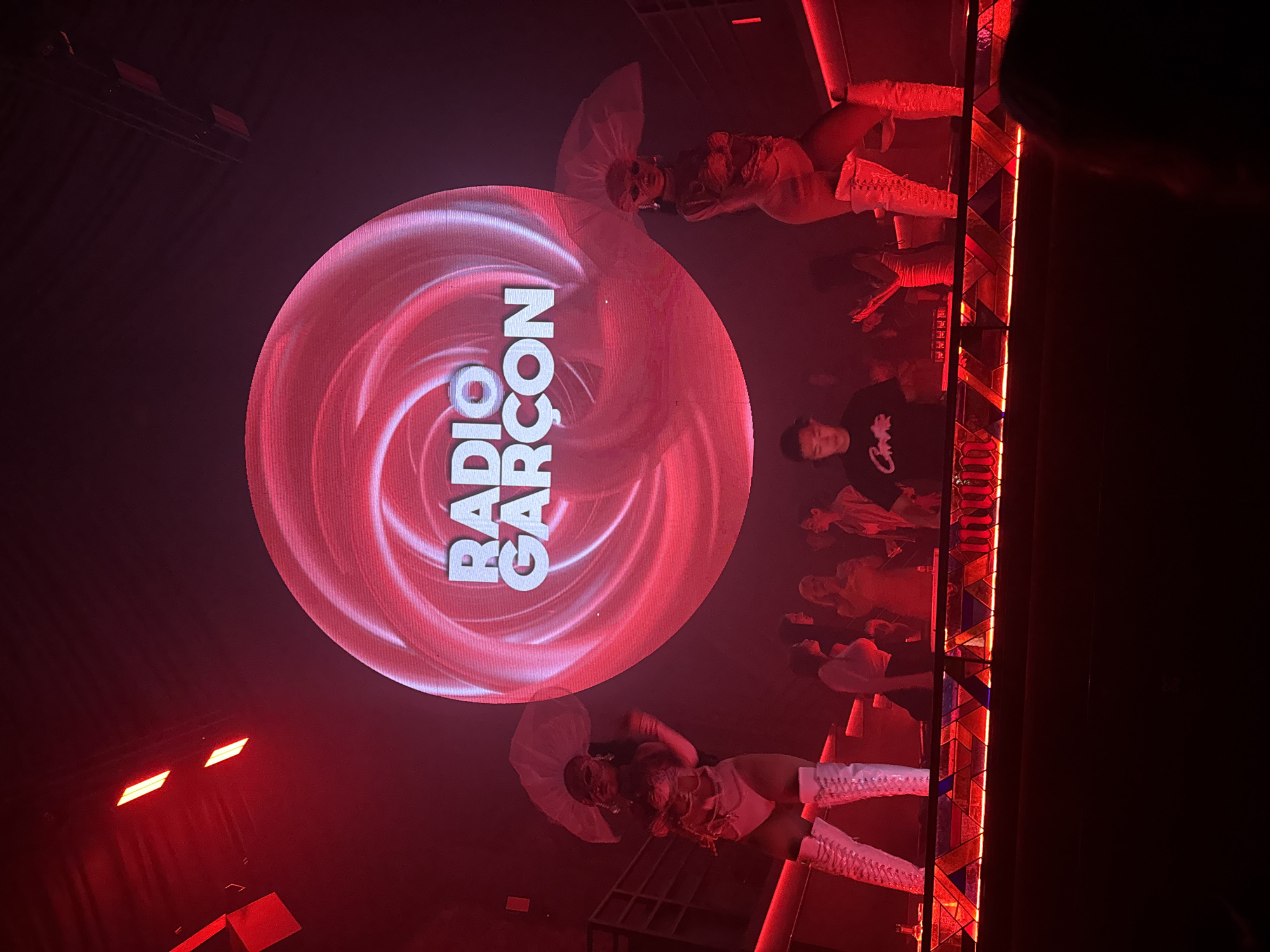 In North Kuta, Indonesia, an indoor club scene is illuminated by deep red lighting. A large, circular screen at the back displays the stylized white text "RADIO GARÇON" within a swirling pink graphic. Two performers, dressed in white, stand on a raised platform to the left and right of the screen. The performer on the left wears a white outfit with a large, translucent headdress and thigh-high white boots. The performer on the right also wears a white outfit with a similar headdress and thigh-high white boots, posing with one hand raised. In front of the performers, on the same platform, a DJ wearing a black t-shirt with white lettering is visible behind a console. A small crowd of people can be seen in the background, some standing and some seated, also bathed in the red light. The platform itself is lit from below with red and blue colored lighting embedded in a patterned glass-like material. The overall ambiance is that of a lively nightclub performance.