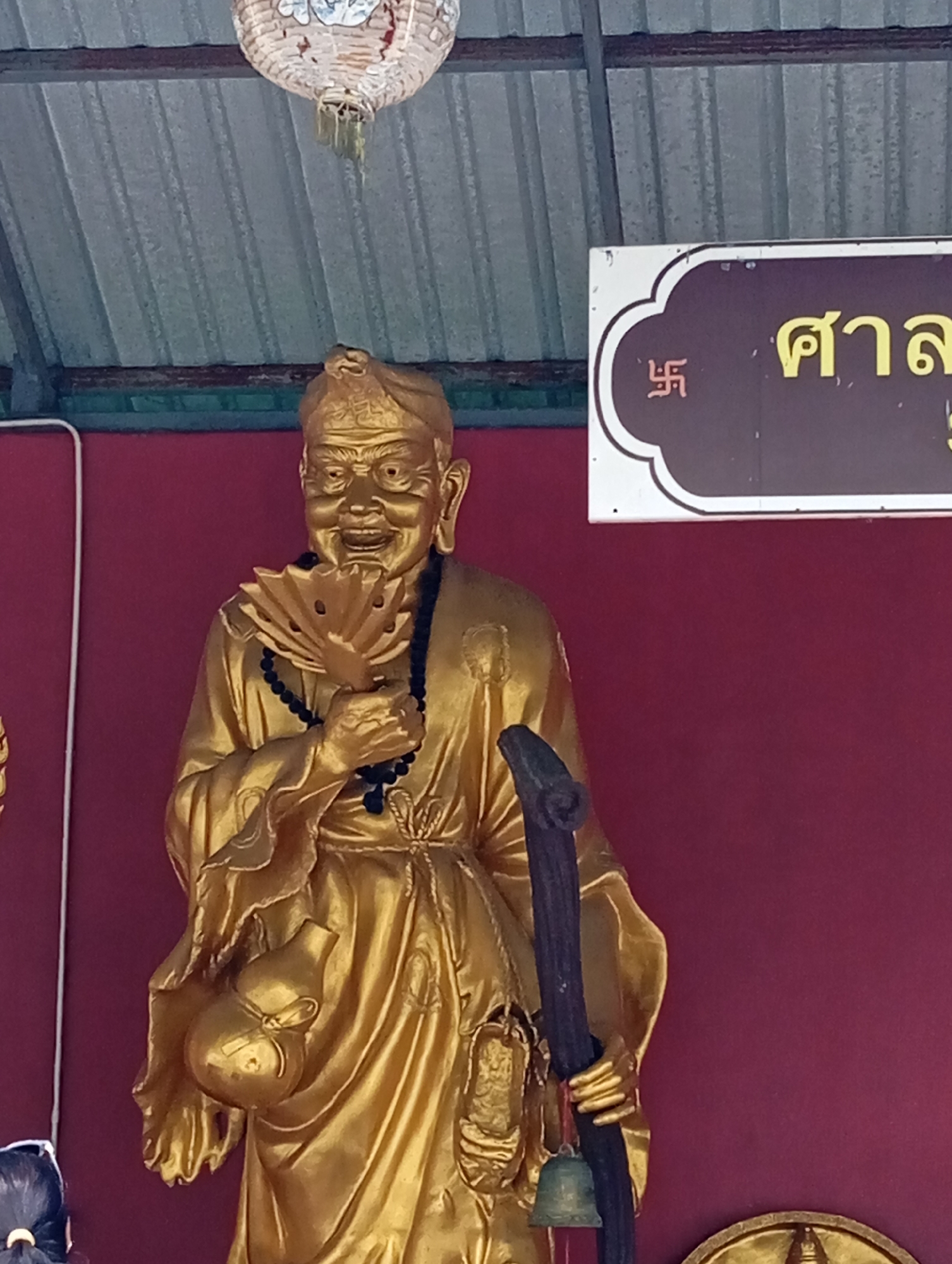 In Karon, Thailand, a golden statue of a man holding a fan stands against a maroon wall. The man wears a robe and a necklace, and a pouch hangs from his waist. He also holds a staff with a bell attached. Above the statue, a lantern hangs from a corrugated metal ceiling. To the right, a sign features Thai script and a swastika symbol.