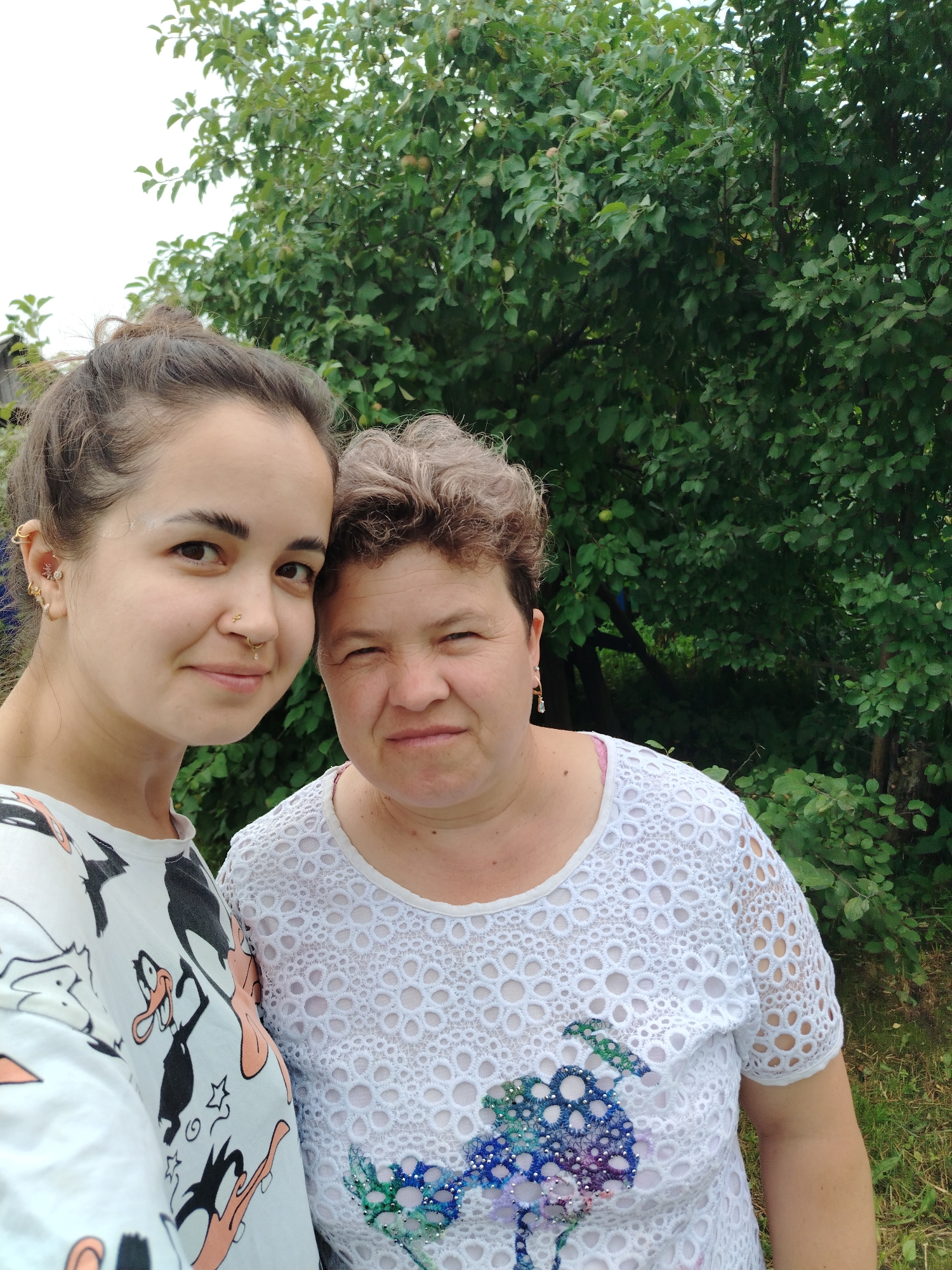Two women are posing for a selfie outdoors, in front of a lush green tree with some small green apples visible. The woman on the left has her hair pulled up in a bun and is wearing a white t-shirt with a cartoon character print. She has several piercings on her left ear and a nose ring. The woman on the right has short, graying hair and is wearing a white lace-like top with colorful floral embroidery. They are both smiling and looking towards the camera. The background suggests a natural, possibly rural setting with foliage and grass. The location is identified as Gorzhan, Russia.