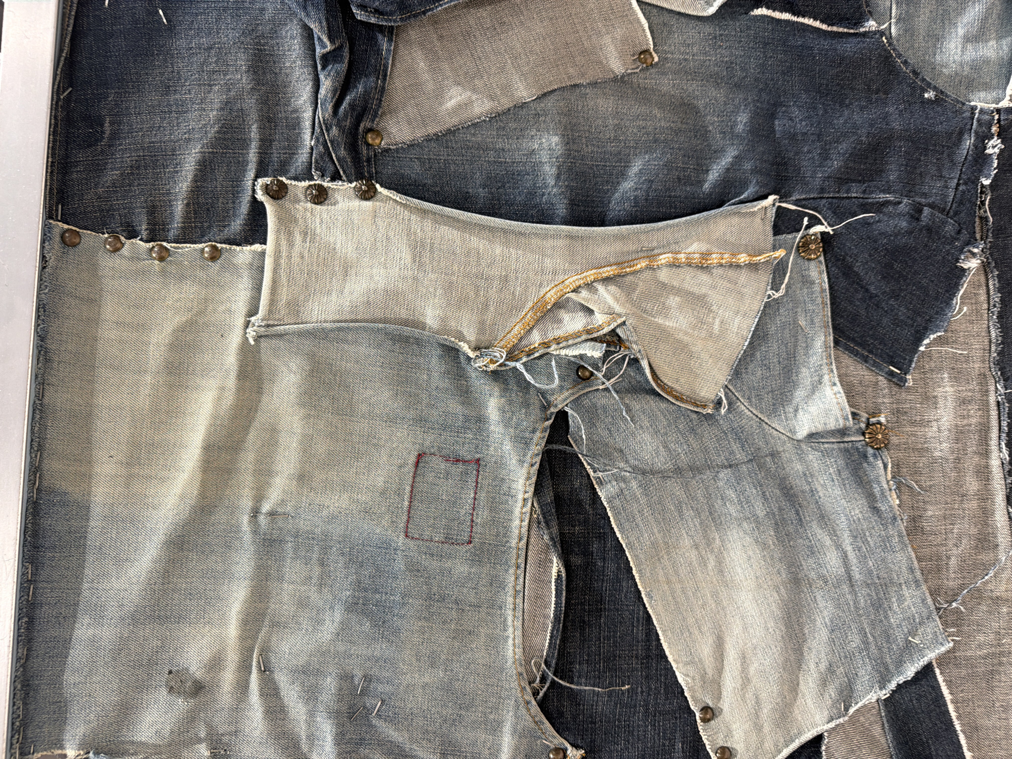 The image is a close-up shot of denim patchwork art, featuring various shades of blue denim, pieced together with metal studs along some of the edges. The denim pieces show wear and tear, with frayed edges and visible seams. A portion of a zipper is visible near the top center of the image. The background consists of more denim patchwork, creating a textured and layered effect. The scene is likely an indoor setting, given the controlled lighting. There's no indication of Spreitenbach, Switzerland, or any specific location. No people are visible.