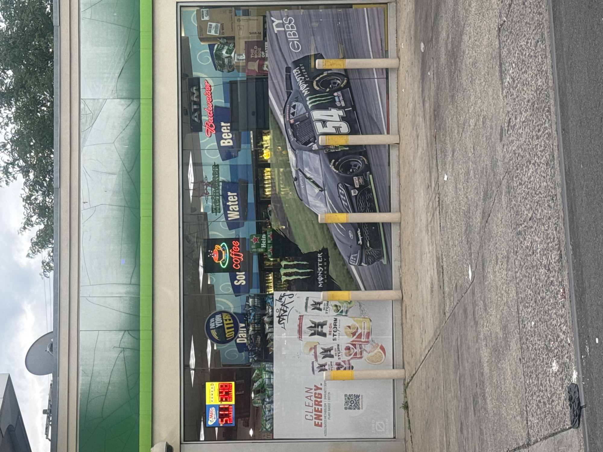 This media file shows a storefront at a convenience store or gas station in New York. The view is from across the street, looking at the large display windows of the store. The store has a green and cream-colored exterior.
Inside the display window, there are numerous advertisements and product displays. Prominently featured is a large poster of a black Monster Energy sponsored NASCAR race car with the number 54. Other advertisements include Budweiser beer, Heineken, Monster Energy drinks, Reign Storm energy drinks, and New York Lottery information. There is also a neon sign for "Sol coffee". Digital displays show lottery jackpot amounts, with one reading "$511 MILLION" for Mega Millions and another "$348 MILLION" for Powerball.
Outside the store, there are several yellow-topped bollards or posts arranged in front of the window. The sidewalk in front of the store is made of grey concrete with visible cracks and some debris. Beyond the sidewalk, a paved road is visible. The weather appears to be overcast, and there is a large tree with green foliage on the left side of the image. The overall impression is a typical urban convenience store environment. No people are visible in the image.
