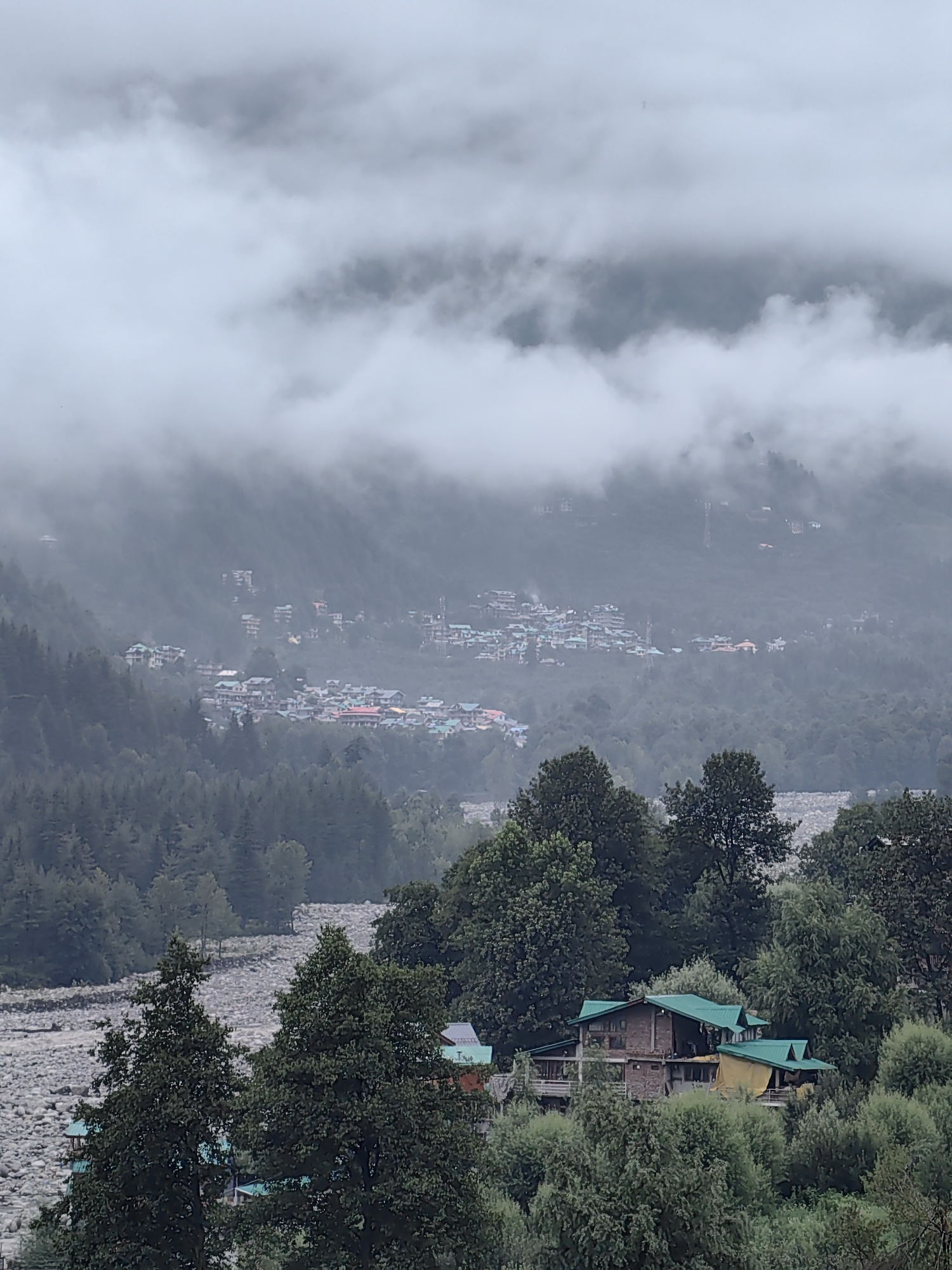 An overcast, misty scene depicts a mountainous region, likely in Vashisht, India. In the foreground, lush green trees partially obscure a cluster of buildings with distinctive green roofs. One prominent structure, possibly a guesthouse or resort, is nestled amongst the foliage. Further in the distance, a village or small town is visible on the side of a hill, characterized by numerous buildings with brightly colored roofs, including red and blue. The town is shrouded in fog, with only the upper portions of the mountains visible through the mist. A rocky riverbed can be seen in the mid-ground, adding to the rugged landscape. The overall atmosphere is serene and cool, suggesting a humid or rainy day.