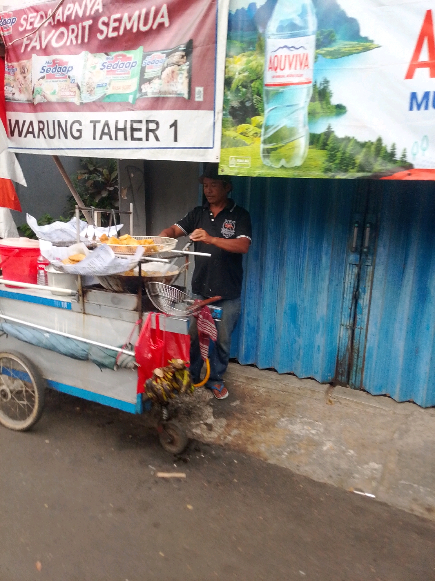 In East Jakarta, Indonesia, a street vendor operates a food cart. The vendor, a man wearing a black t-shirt with a white collar and blue jeans, stands beside his cart, which is laden with fried foods, such as bananas and what appear to be spring rolls. The cart itself is a mobile unit with wheels, a metal frame, and a blue and white exterior. A large banner hangs above the cart, displaying text in Indonesian related to food and the vendor's establishment, "WARUNG TAHER 1". Another banner to the right features an advertisement for a bottled water brand, AQuaviva, against a backdrop of a scenic landscape. The environment is urban, with a paved street in the foreground and a corrugated blue metal wall forming the background, suggesting a shop or market stall. The scene is captured during daylight.