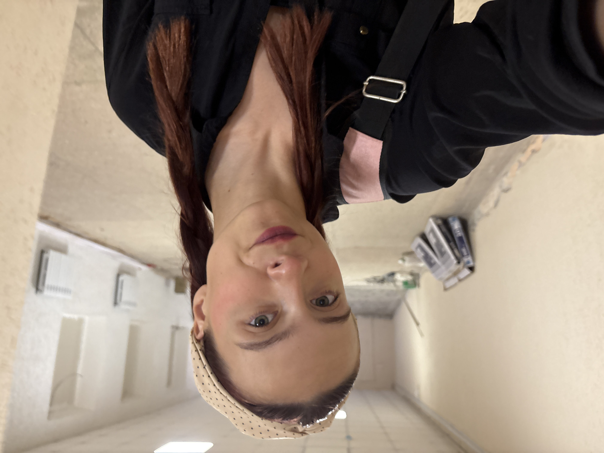 A young woman with reddish-brown hair pulled into two braids and wearing a tan polka-dot headband and a black shirt with a pink accent is taking a selfie in a hallway. The hallway appears to be under renovation, with exposed walls, some construction materials leaning against the wall, and visible heating radiators. The lighting suggests it may be daytime, with artificial lights on in the ceiling. The background is blurred, indicating a shallow depth of field. The location is identified as Chaikovsky, Russia.