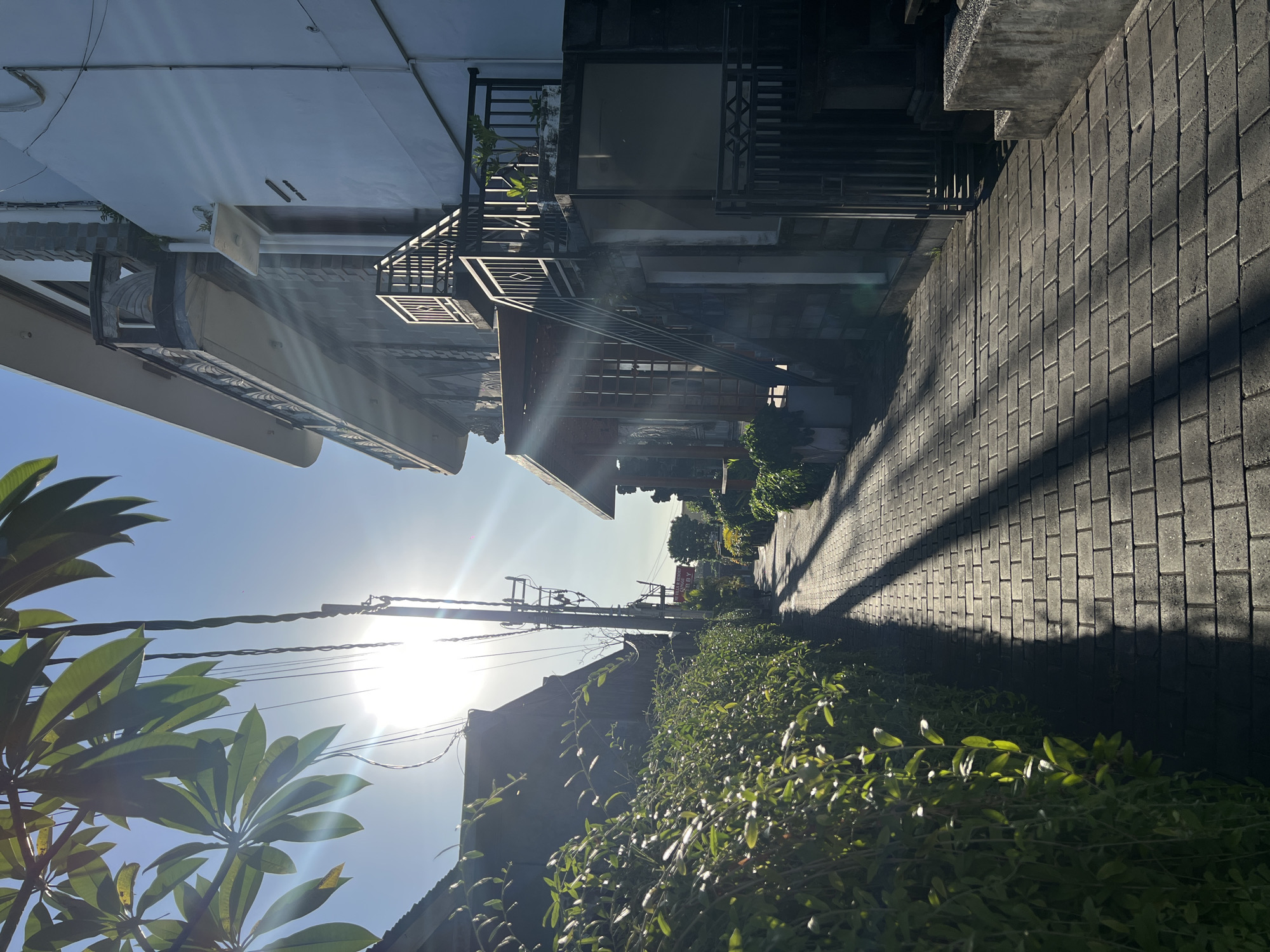 A sun-drenched alleyway in North Kuta, Indonesia, is seen from a low angle. The narrow path is paved with gray brick and runs between buildings and greenery. Sunlight casts sharp shadows across the brickwork. A building to the left features external stairs and decorative metalwork. Lush green foliage lines the bottom right of the frame, contrasting with the hard lines of the architecture. Overhead, a clear blue sky is pierced by the bright sun, with utility wires cutting across the frame.