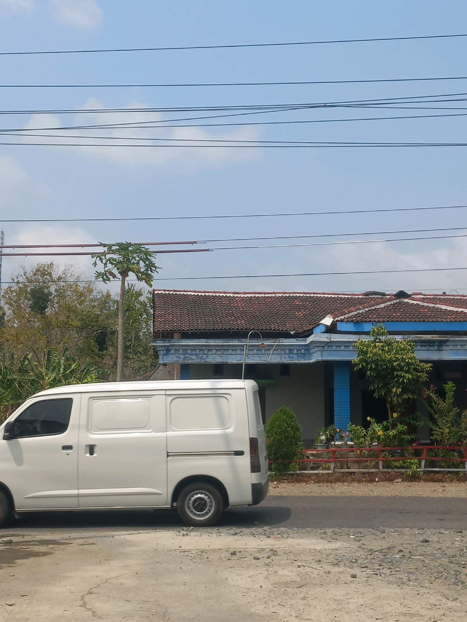 A white van is parked on the side of a road in Baturetno, Indonesia. The van is positioned to the left of the frame, with its side facing the viewer. Behind the van is a house with a red tiled roof and blue accents. The house has a covered porch area with decorative trim. In front of the house, there is a low red fence and various plants and small trees. A tall, slender tree with green foliage stands near the center of the image, between the van and the house. Power lines are visible crisscrossing the sky above the scene. The sky is partly cloudy with a light blue hue. The ground in the foreground is a mixture of dirt and gravel.