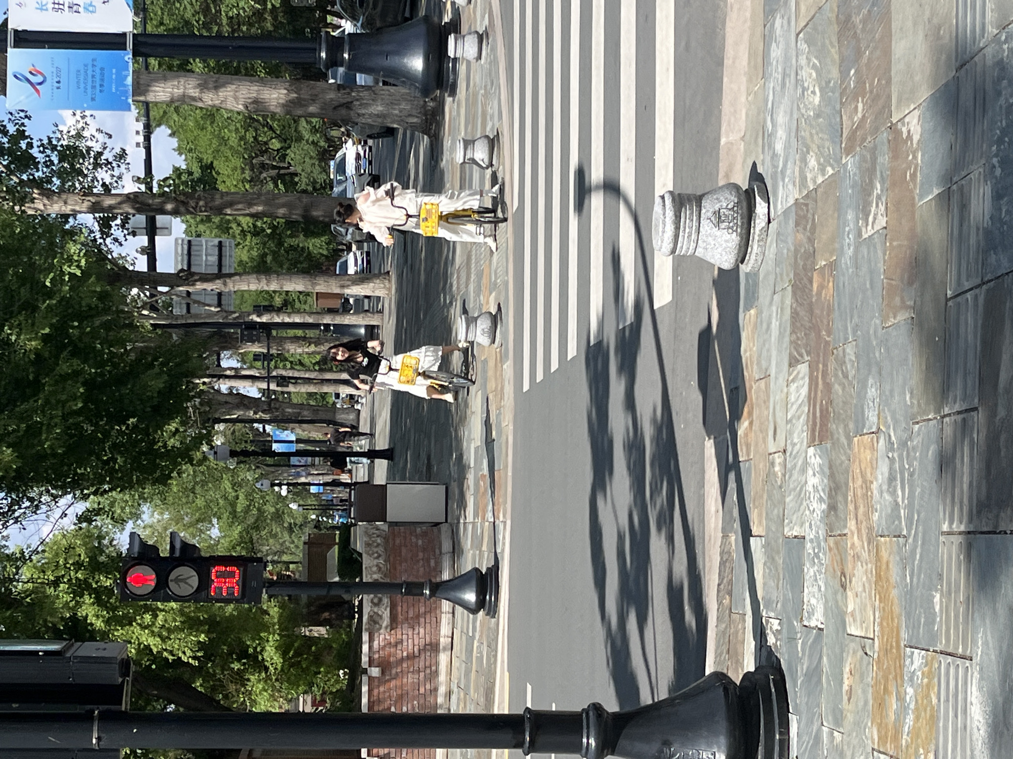 The image depicts a street scene in Changchun City, China. Two women are riding bicycles across a crosswalk. The traffic light shows a red "walk" signal. There are trees lining the street and a banner with Chinese characters is visible. The street is paved with stone tiles. Black bollards are installed as barriers for pedestrians.