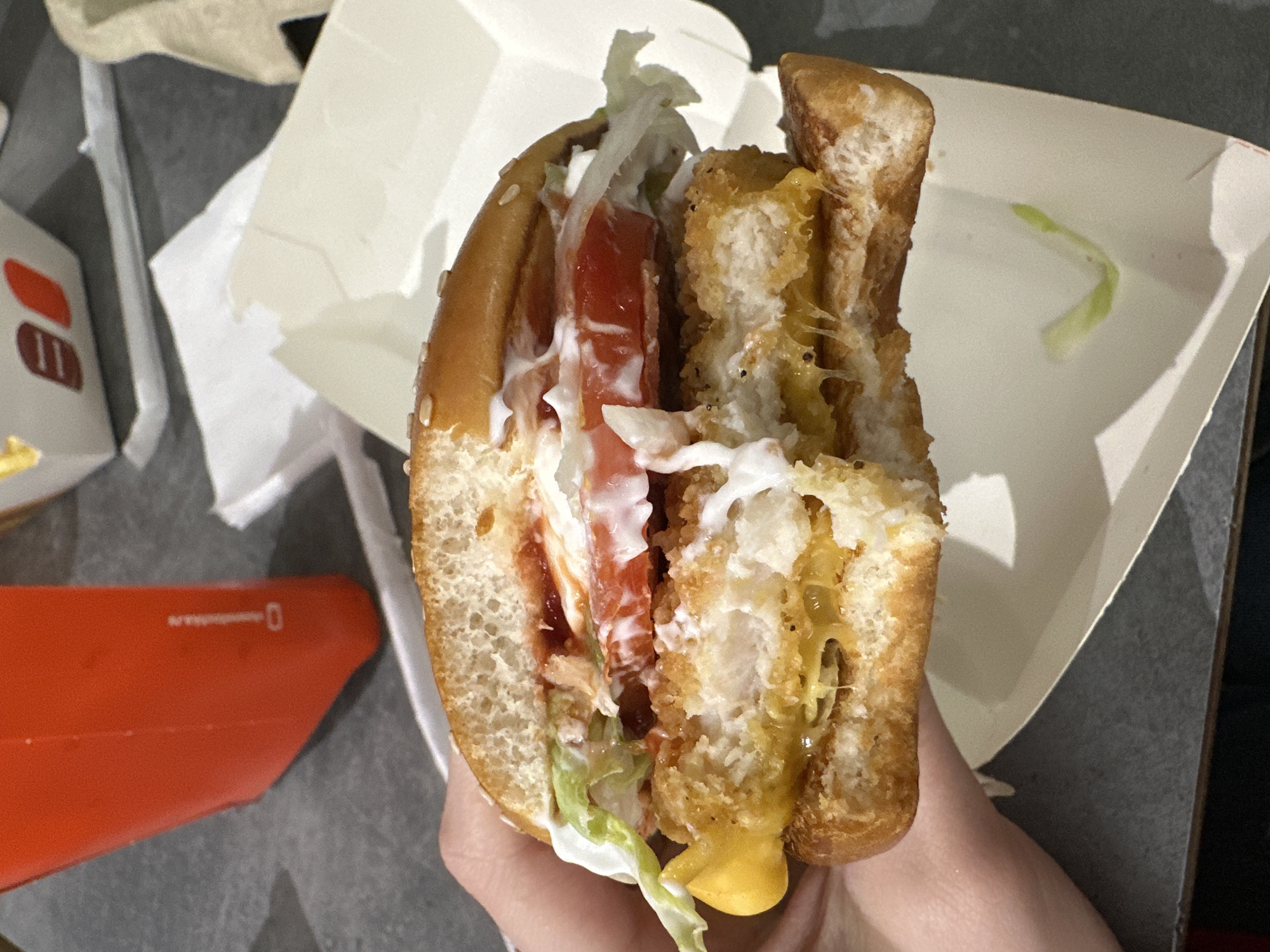 A close-up image depicts a partially eaten chicken sandwich, likely a Zinger Cheese sandwich from the KFC chain, held in a person's hand. The sandwich consists of a sesame seed bun, fried chicken patty, cheese, lettuce, tomato, and sauces. The bun has been torn in half revealing the sandwich's interior. The sandwich sits in a white cardboard container. A discarded red container with a logo and a white paper wrapper are visible in the background. Based on other information, the image was taken in Kazan, Russia.