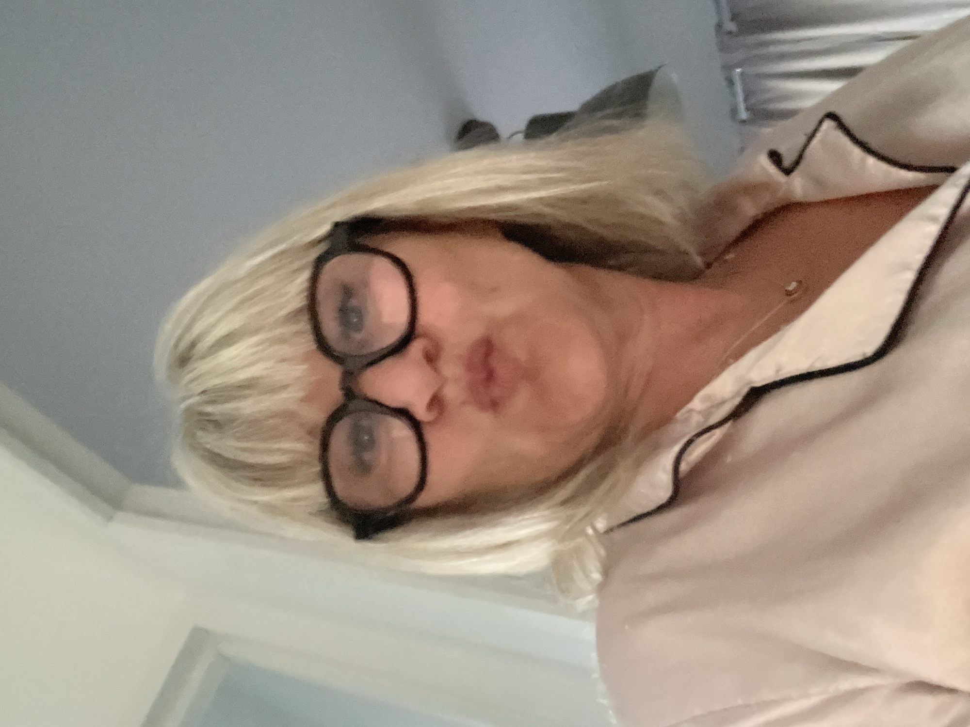 An eye-level selfie shows a person with short, light blonde hair and black-framed glasses, making a pursed-lip expression. The person is angled upwards towards the camera, which is positioned below their chin. They are wearing a light-colored (possibly cream or pale pink) pajama-style top with black piping detail along the collar and lapel. A delicate gold-colored necklace with a small pendant is visible around their neck. The background is a plain, light grey or off-white wall, suggesting an indoor setting. Reflections in the glasses show a circular light source. The image is captured in a vertical orientation, but the subject's head is tilted to the left, appearing rotated. There is no visible text. The scene is set in Chambœuf, France.