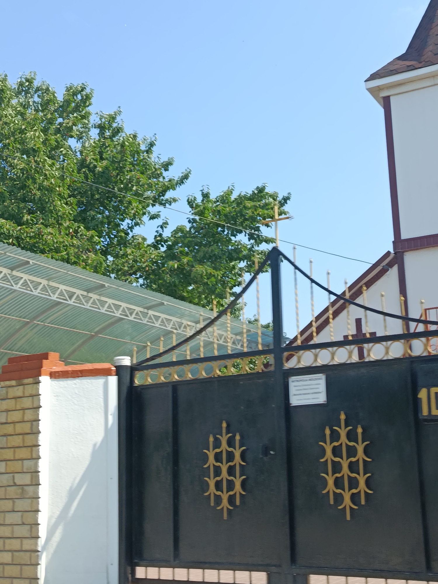 The image depicts a black metal gate with ornate golden scrollwork, set against a clear blue sky. To the left of the gate, a white pillar is visible, partially covered by a light-colored brick wall. Behind the gate and pillar, a transparent, corrugated metal roof structure is present, supported by a white metal frame. Lush green trees with dense foliage are seen behind the roof structure. In the background, a white building with a brown-tiled roof and dark trim is partially visible, topped with a golden cross. Another smaller golden cross is affixed to the building's facade. A sign is attached to the gate, and the number "10" is visible on the right side of the gate. The scene appears to be an exterior view of a church or religious institution, possibly located in Chișinău, Moldova.