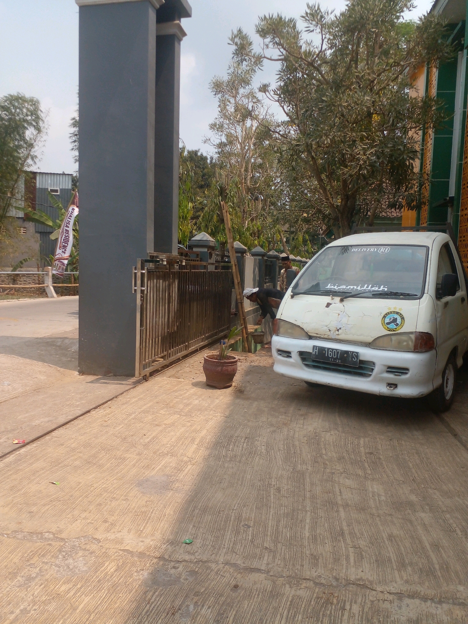 In Banyuputih, Indonesia, a white van with license plate H-1607-YS is parked in front of a grey pillar and a metal gate. The van has "DELIVERY" written on its windshield and a sticker with an emblem on the passenger side. Two individuals are visible near the van. One person, wearing a white cap and dark clothing, is leaning over a bucket next to the gate. Another person, wearing a patterned shirt and a cap, stands behind them. A red flower pot sits on the concrete ground to the right of the gate. In the background, trees and some buildings are visible, including a structure with green and yellow decorative elements. A white banner with red text that reads "KHALISA RESIDENCE" hangs from a pole on the left side of the image. The ground is a textured concrete surface.