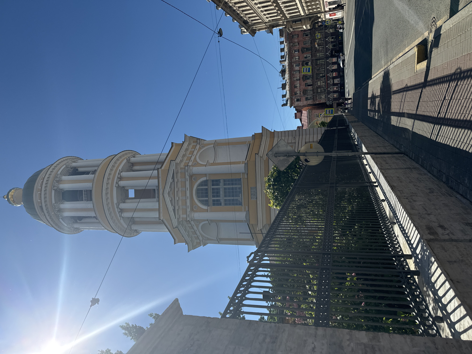The image, oriented vertically, depicts a street scene in Saint Petersburg, Russia. The primary subject is a tall, ornate bell tower, painted in pale yellow and white, with a domed roof. The tower features multiple levels with columns and arched windows. To the right of the tower, a street with a sidewalk is visible, bordered by a black metal fence. Beyond the street, buildings with multiple stories and windows line the road.
The time of day appears to be daytime, indicated by the bright sunlight and distinct shadows cast on the sidewalk and fence. The sky is a clear, bright blue, suggesting good weather. The scene is likely an urban environment, given the presence of buildings, a street, and pedestrian infrastructure like sidewalks and a road.
There are no people clearly visible in the image, though distant figures might be present in the background. The focus is on the architecture and the streetscape. Notable details include the intricate design of the bell tower, the patterned shadows from the fence and buildings, and the overall sunny atmosphere.
Some visible text includes pedestrian crossing signs with yellow triangles and black symbols. One circular road sign with a diagonal line is also present near the tower. The image evokes a sense of urban exploration and architectural appreciation.