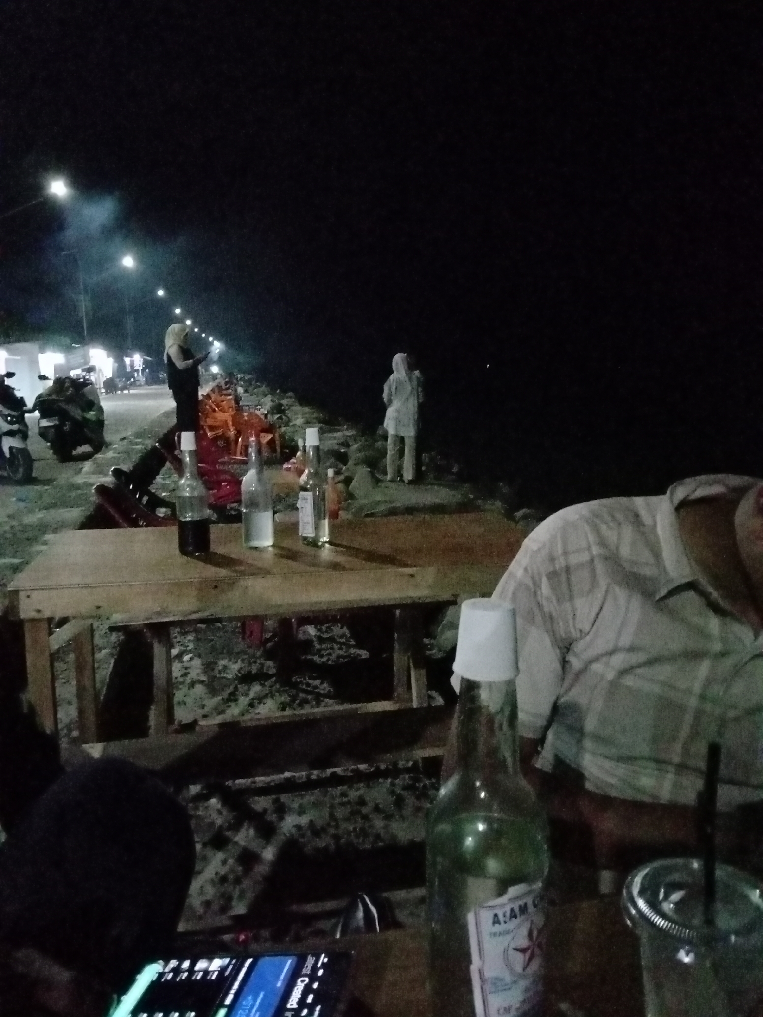 The image shows an outdoor nighttime scene in Lhokseumawe, Indonesia. A wooden table is in the foreground, cluttered with several open bottles, a plastic cup with a straw, and a smartphone displaying an interface. A person wearing a plaid shirt is seated at the table. In the background, two people wearing headscarves stand near the water's edge. Streetlights are visible in the distance, illuminating an area where motorbikes are parked. The overall scene suggests a casual gathering in a dimly lit outdoor setting.