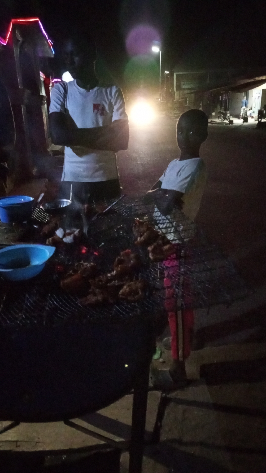 Here's a forensic description of the scene:
**Location:** Sabon Tasha, Nigeria.
**Environment:** The scene is set at night. It appears to be on a street, with buildings visible in the background. The lighting is dim, with the primary light source being a street lamp. There's also a bright light from an approaching vehicle (likely a car or motorcycle) in the distance.
**Subjects:** Two people are present, an adult and a child.
* The adult stands behind a grill. The adult has their arms folded.
* The child is next to the grill.
**Objects:**
* A large charcoal grill is present, covered with a metal grate.
* Pieces of meat are on the grill, seemingly being cooked. There is visible smoke and heat emanating from the food.
* Two blue bowls are on the table.
* A small silver pan on the grill.
* A metal grill utensil is visible.
**Actions & Interactions:** Food preparation is taking place on the grill. The adult and child appear to be observing the cooking process.