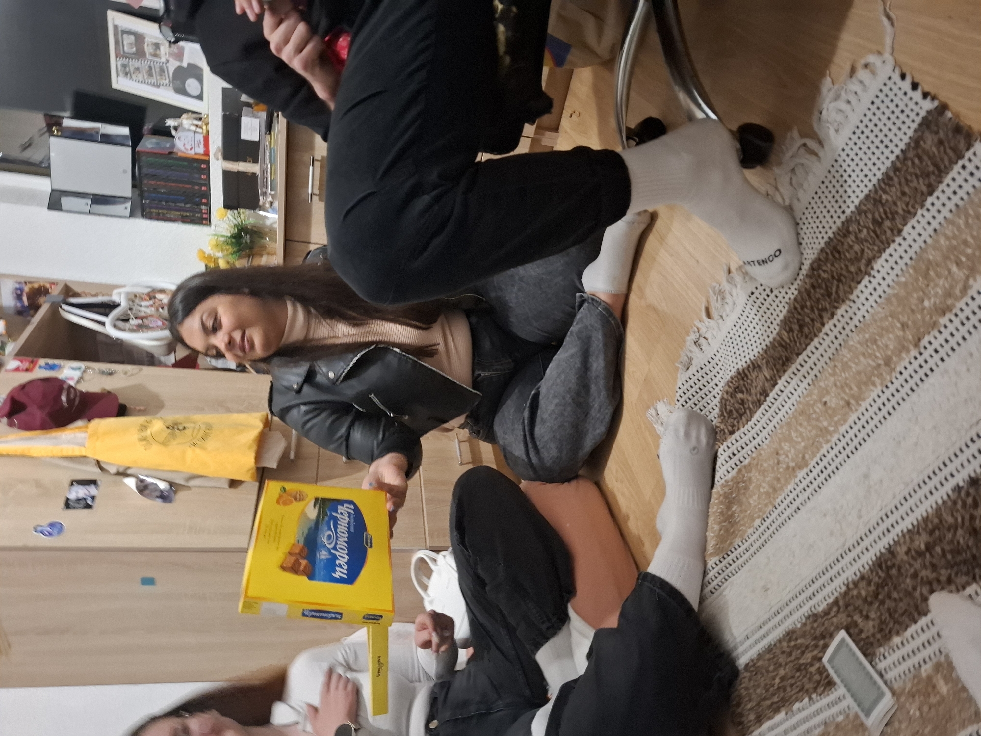 The image shows an indoor scene, likely a home interior in Panagyurishte, Bulgaria. Three people are present, casually seated on the floor. One person, wearing a black leather jacket and grey jeans, is holding a yellow box of "ЧЕРНОМОРЕЦ" candies. The other two people are dressed in similar casual attire, with at least two wearing white socks. The setting includes a striped rug, wooden flooring, and shelves stocked with items in the background. The interaction seems informal, suggesting a relaxed gathering.