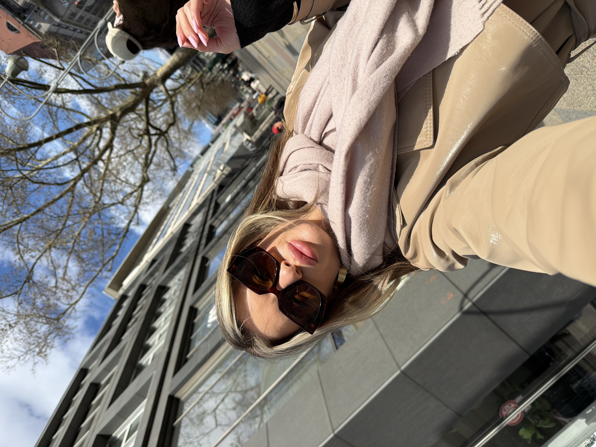 A person is taking a selfie in Berlin, Germany, with a building visible in the background. The individual is wearing sunglasses, a scarf, and a tan jacket. Their nails are painted pink, and they are holding the camera up at an angle. The background includes a bare tree, a modern building with black and white rectangular designs, and a partial view of the sky with clouds. The ground is grey and appears to be a sidewalk or paved area.
